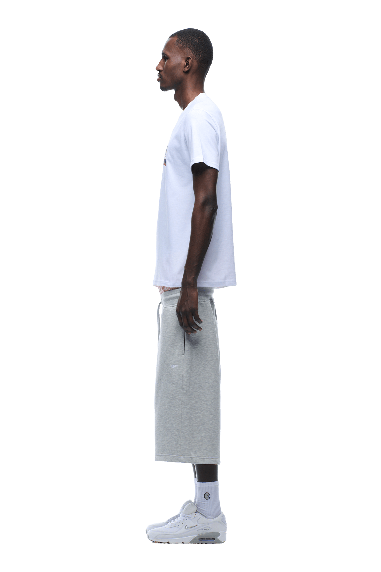 A man stands in profile wearing the 6PM CLEAN EDGE WAVE TEE in white, gray knee-length shorts, white crew socks, and white sneakers against a plain background.