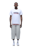 A man faces forward, wearing the 6PM CLEAN EDGE WAVE TEE in white over a striped shirt, paired with loose light gray knee-length shorts and white sneakers. The background is transparent.
