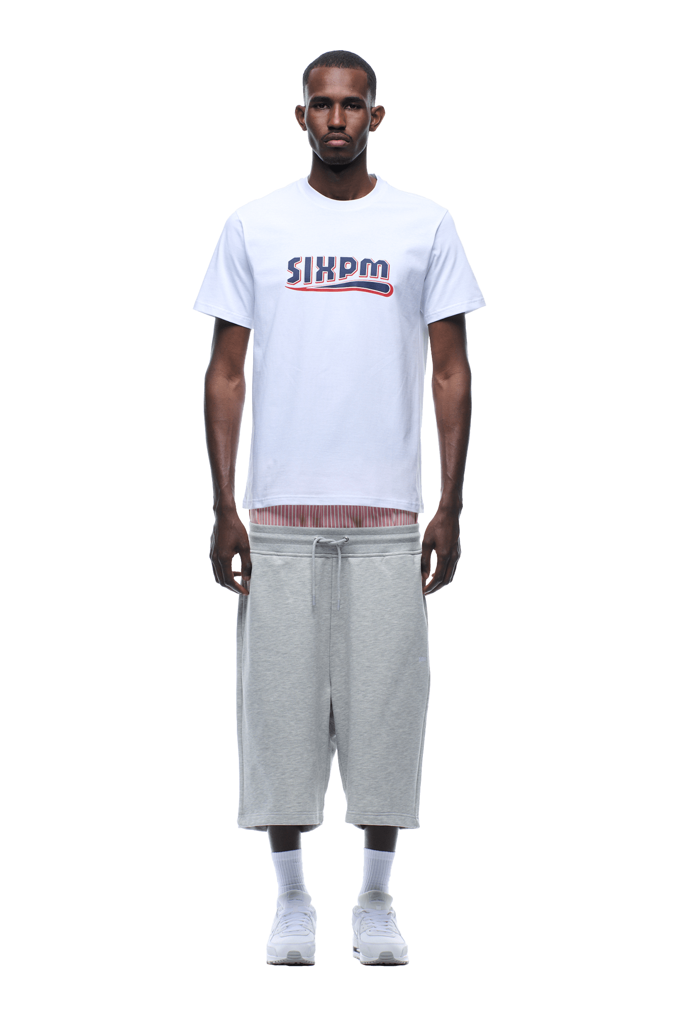 A man faces forward, wearing the 6PM CLEAN EDGE WAVE TEE in white over a striped shirt, paired with loose light gray knee-length shorts and white sneakers. The background is transparent.