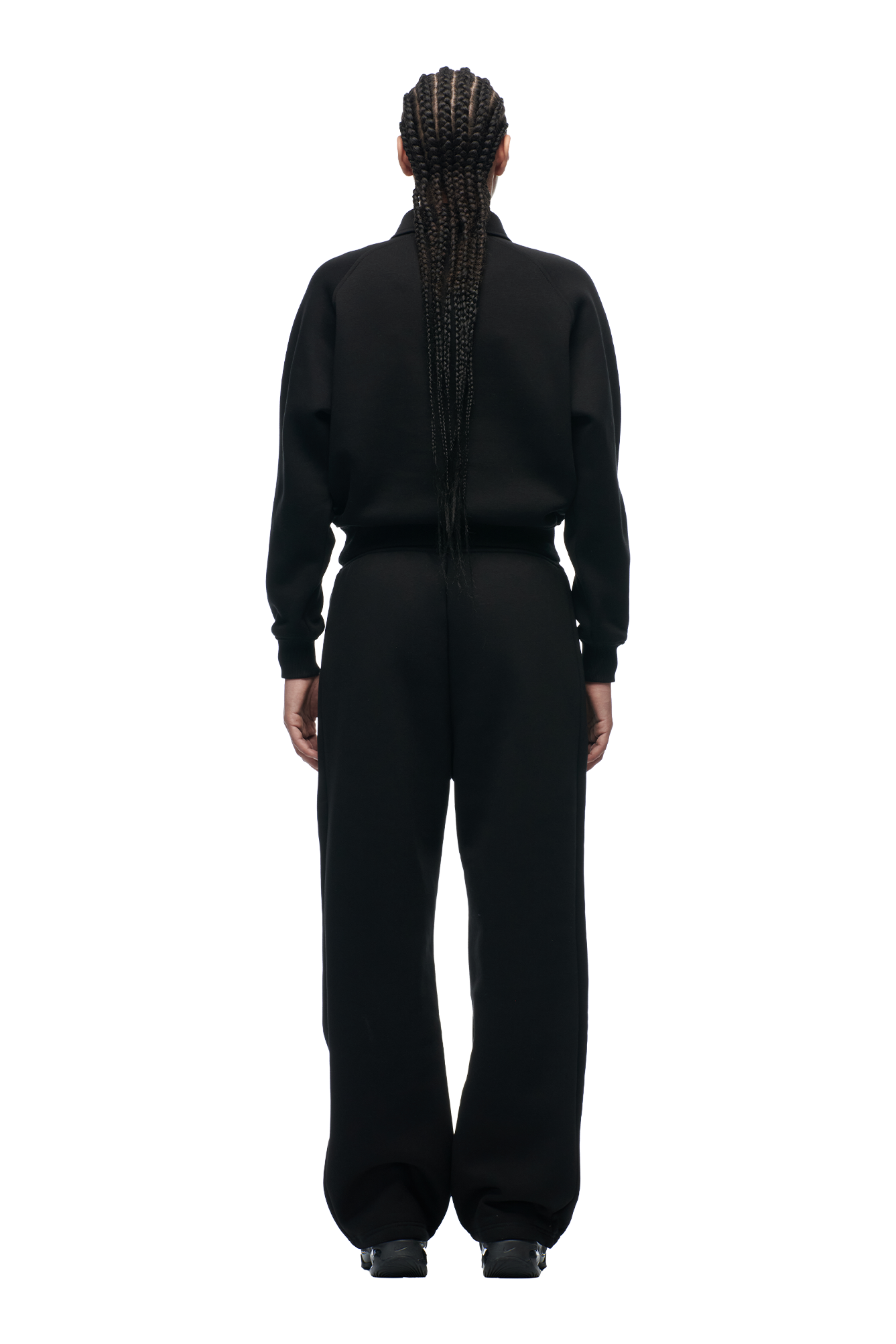 A person with long, braided hair stands facing away from the camera, wearing the 6PM WMNS POLO BLACK long-sleeve top and matching black pants. The background is plain white.