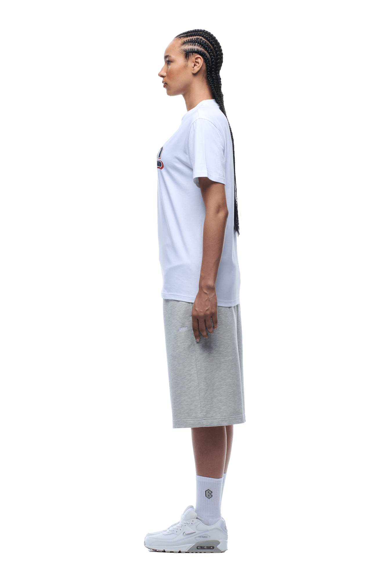 A woman with long braided hair stands in profile, wearing the 6PM CLEAN EDGE WAVE TEE WHITE, light gray knee-length shorts, white socks, and white sneakers.