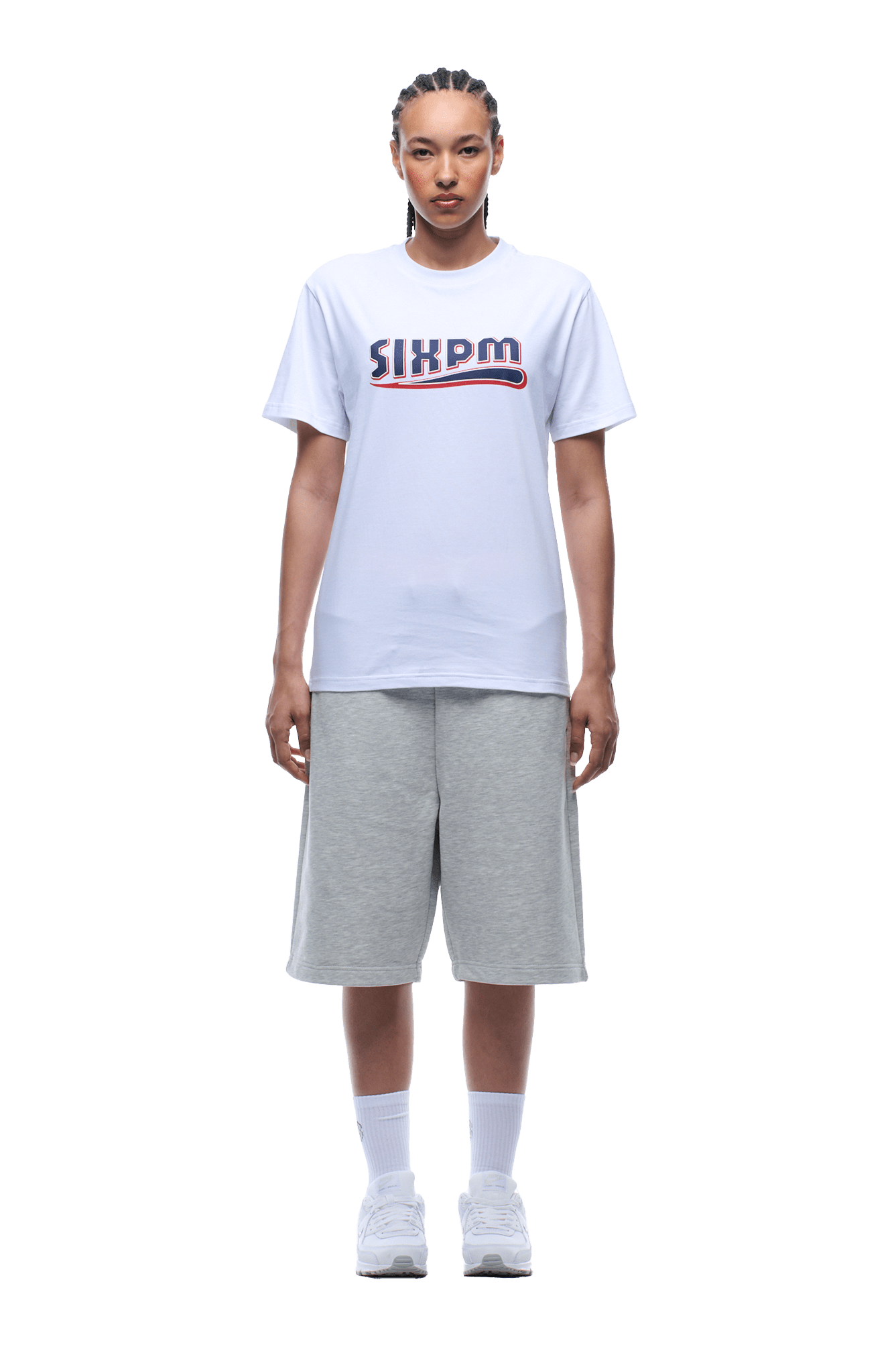 A person with braided hair and a neutral expression faces forward, wearing a 6PM CLEAN EDGE WAVE TEE WHITE, light gray knee-length shorts, white crew socks, and white sneakers.