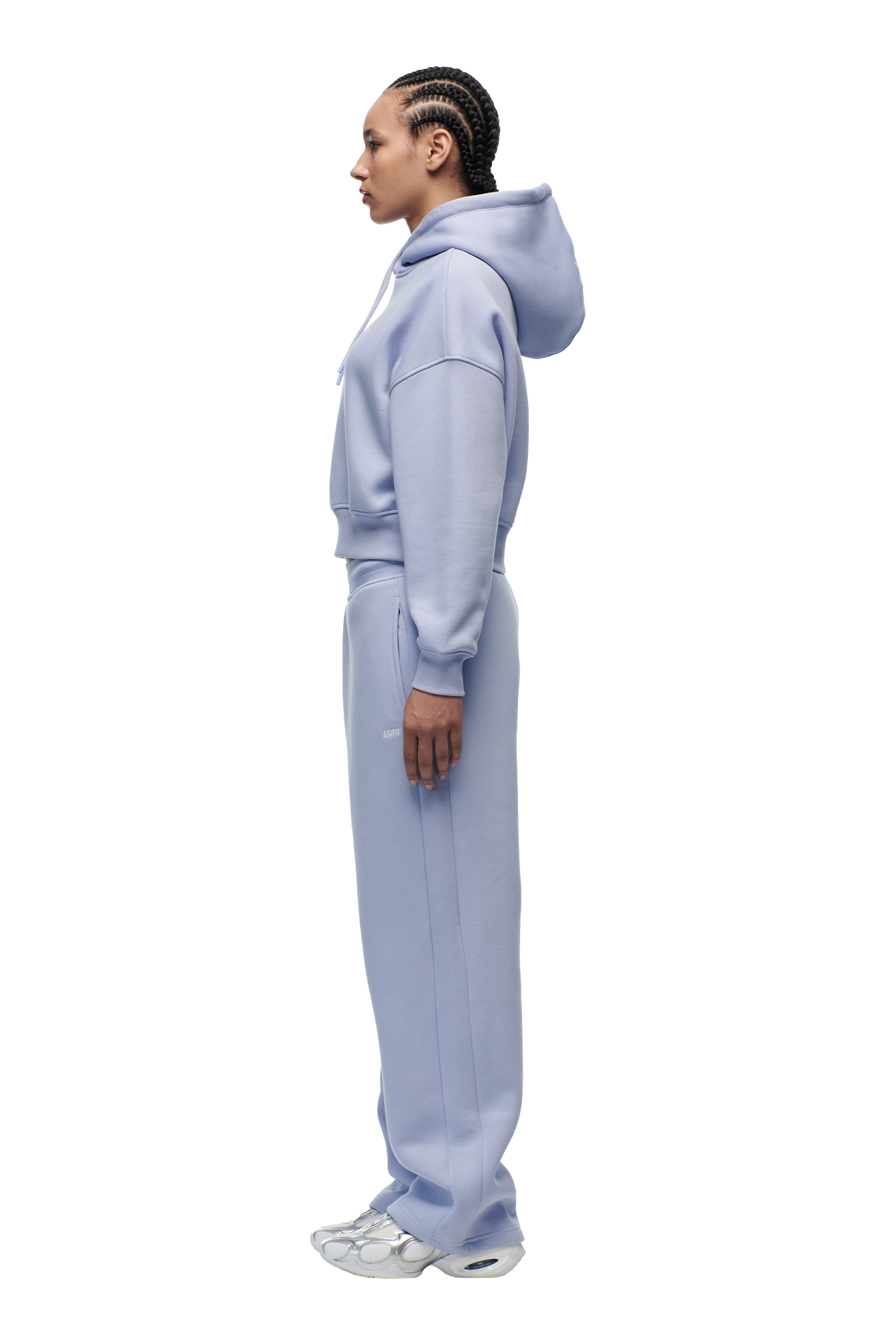 A person with braided hair stands in profile, wearing the 6PM WMNS CROPPED HOODIE SOFT VIOLET and matching sweatpants with white sneakers, set against a plain white background.