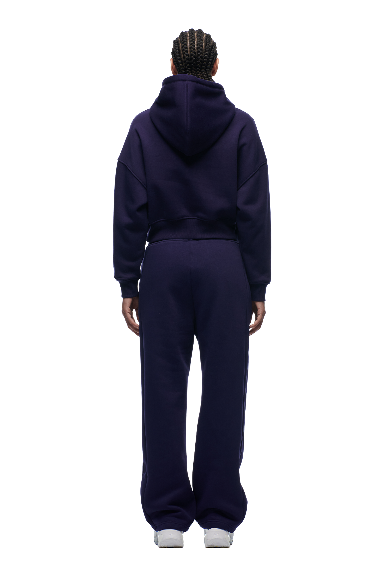 A person with braided hair faces away, wearing the 6PM WMNS Cropped Hoodie in Orbit Blue and matching sweatpants with white sneakers, set against a plain white background.