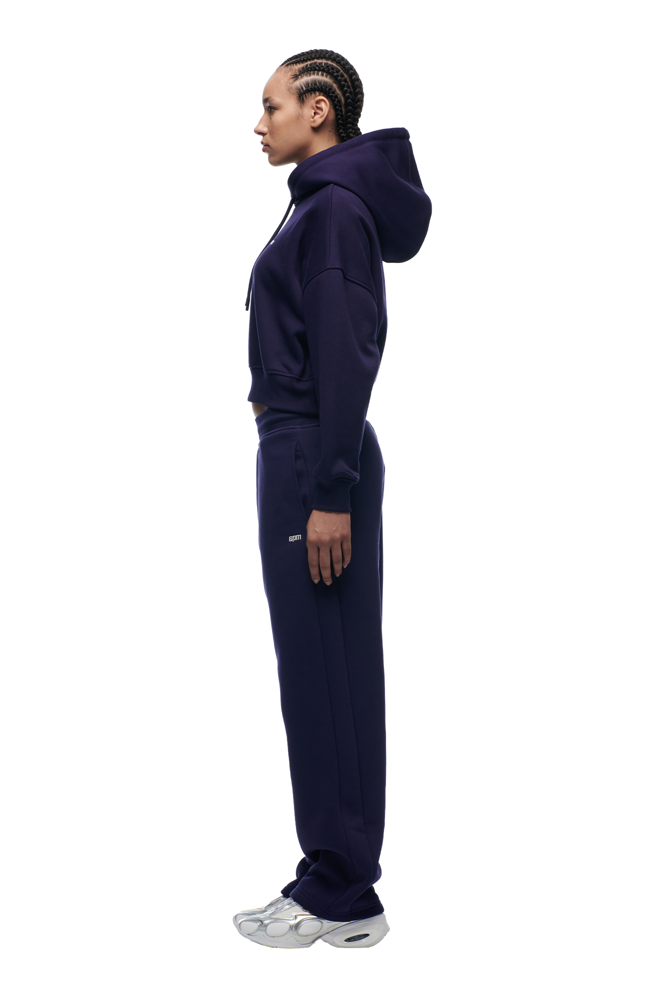 A person with braided hair stands in profile against a white background, wearing the 6PM WMNS Cropped Hoodie in Orbit Blue, paired with matching sweatpants and silver-white sneakers.