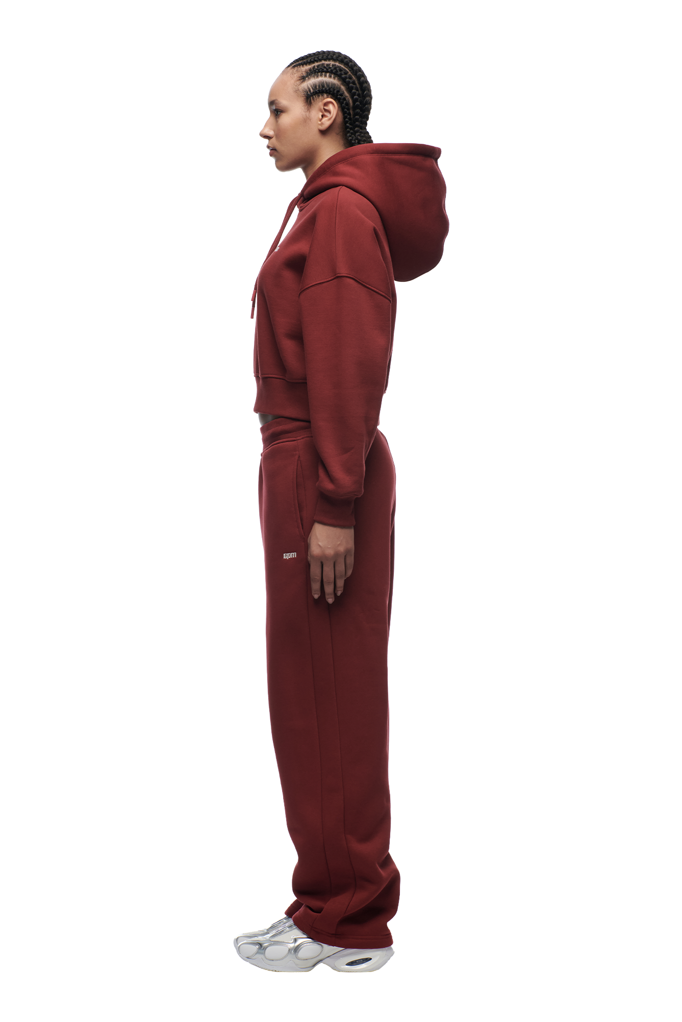A person with braided hair stands in profile against a white background, wearing the 6PM WMNS Cropped Hoodie in Wine Red paired with matching sweatpants and white sneakers.