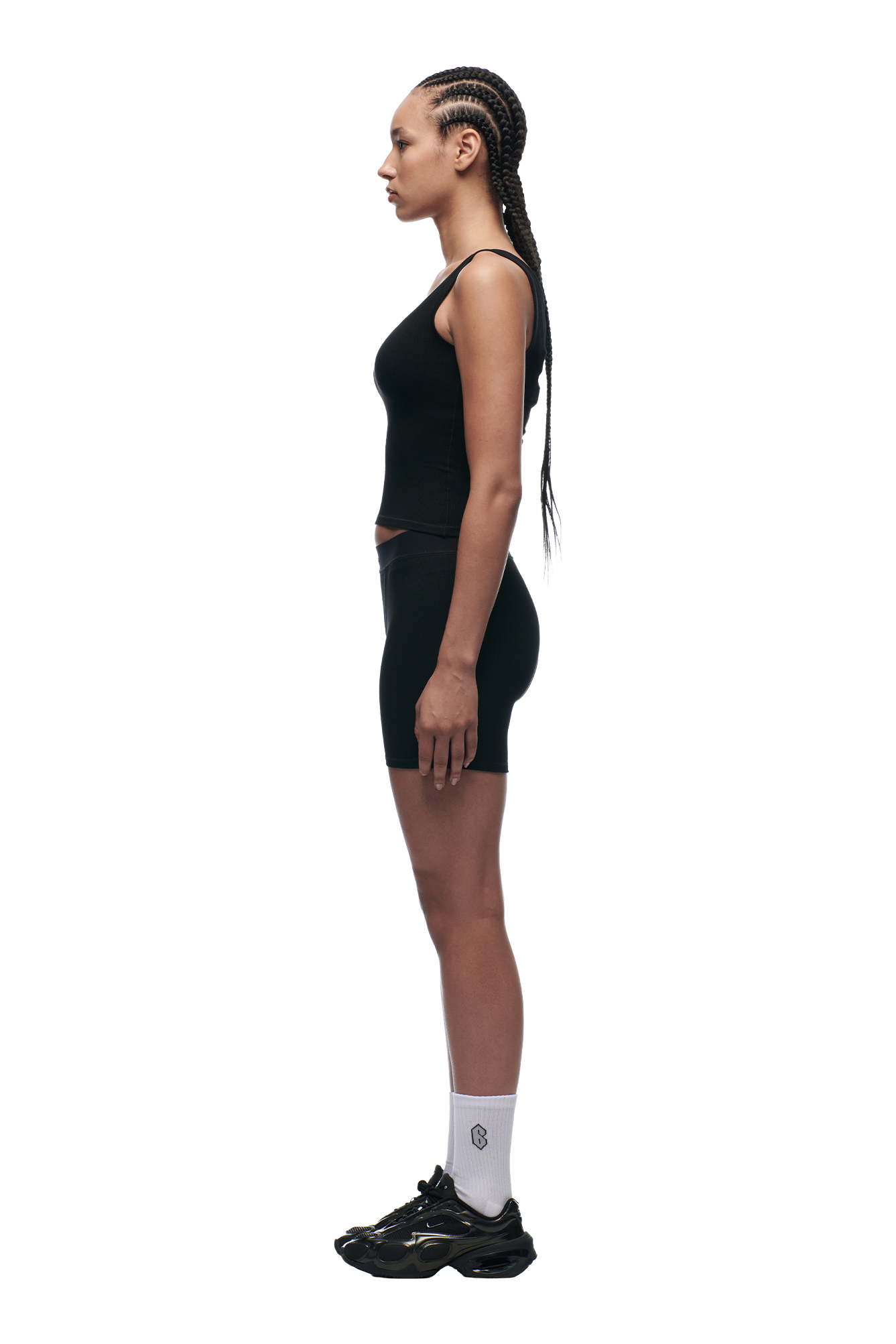 A person with long braided hair stands in profile, wearing 6PM WMNS BOXER SHORTS BLACK, a black tank top, white socks, and black sneakers against a plain white background.