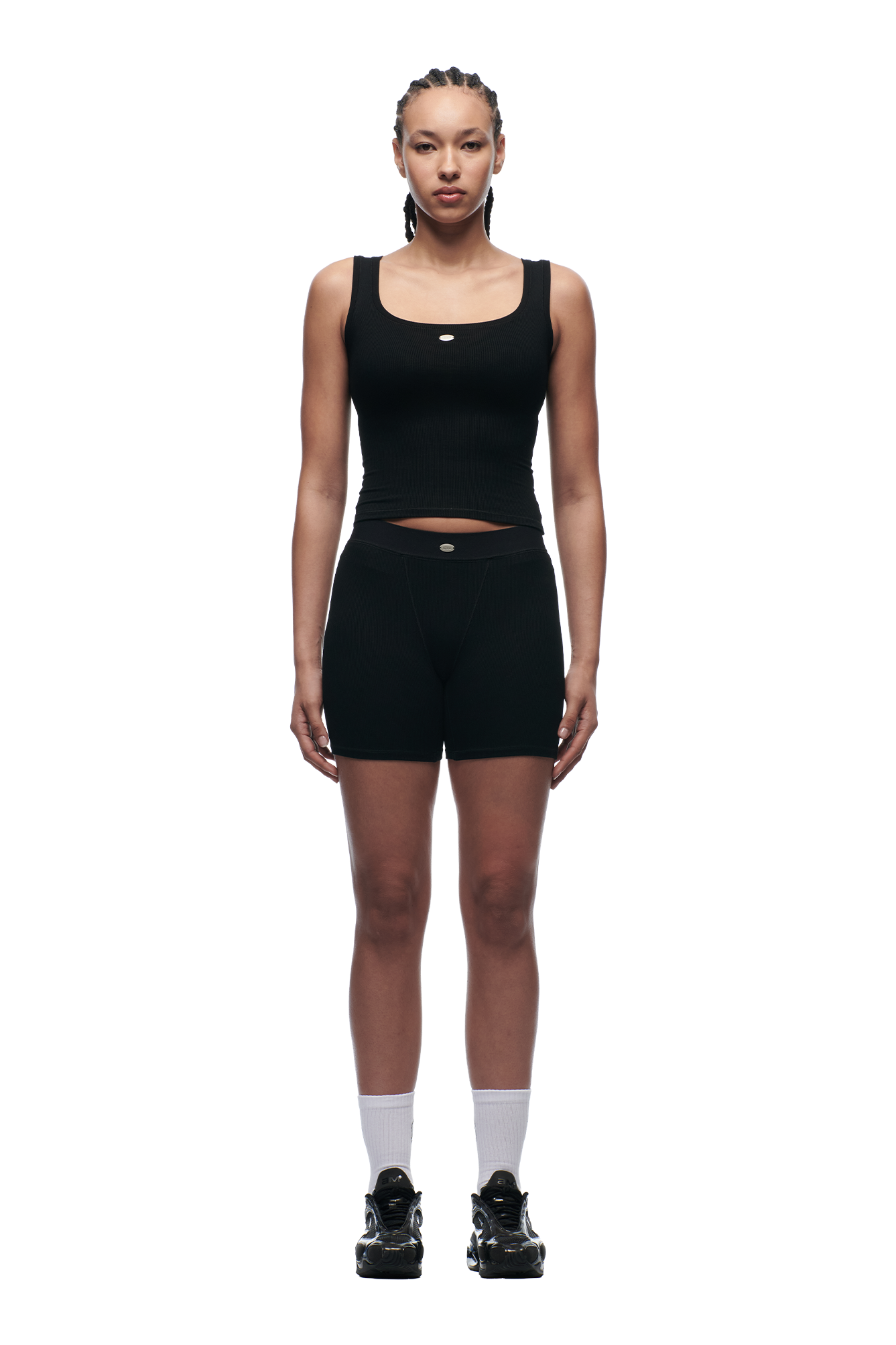 A person with cornrow braids stands facing forward against a white background, wearing the 6PM WMNS TANKTOP BLACK, black shorts, white socks, and black sneakers.