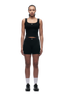 A person with braided hair stands facing forward, wearing 6PM WMNS BOXER SHORTS BLACK, a black tank top, white socks, and black sneakers against a plain white background.