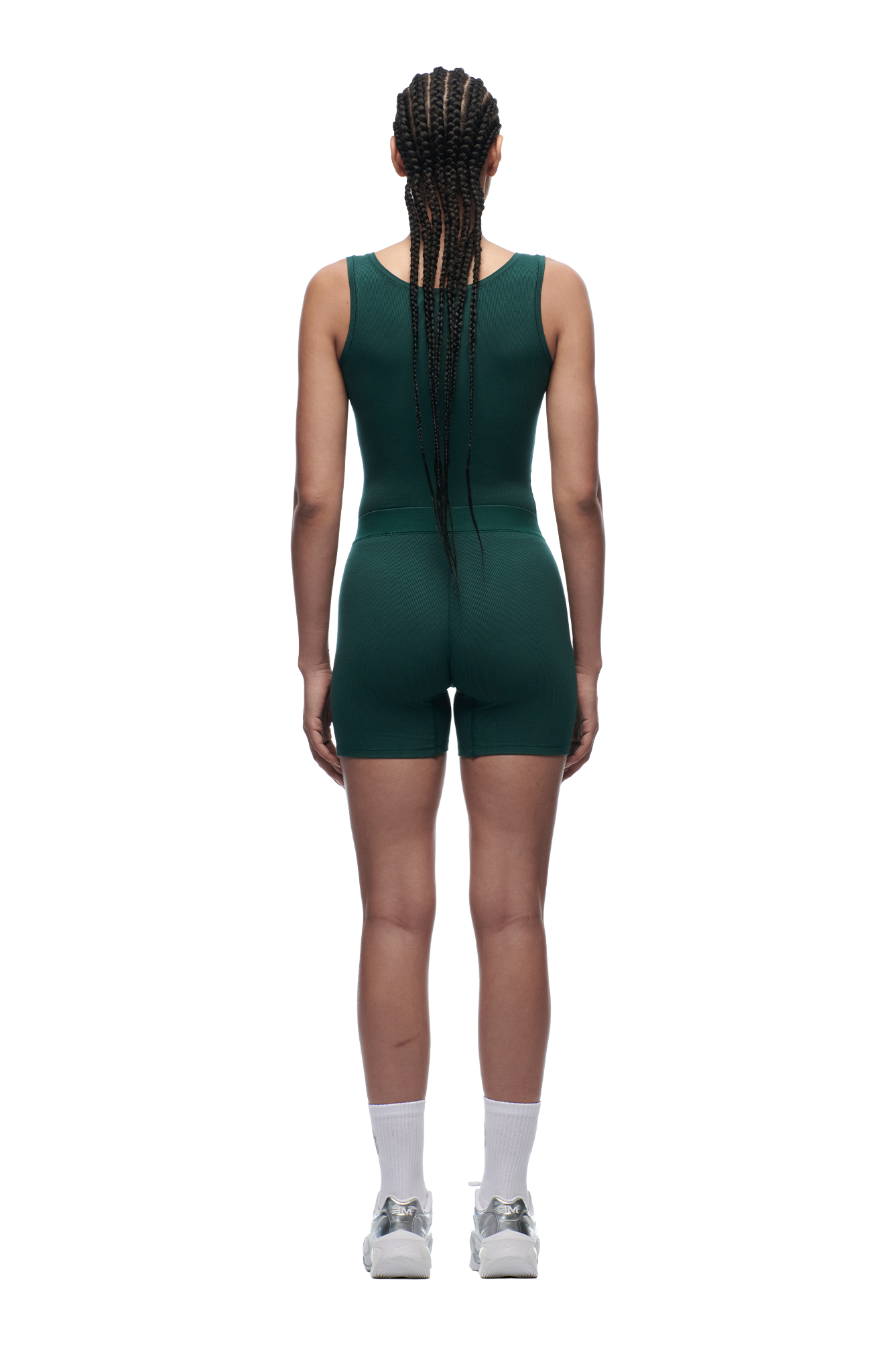 A woman with long braids stands with her back to the camera, wearing 6PM's WMNS BOXER SHORTS EVERGREEN romper paired with white crew socks and silver athletic shoes, set against a plain white background.