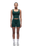 A woman with braided hair faces forward wearing 6PM WMNS BOXER SHORTS EVERGREEN, a green fitted tank and high-waisted shorts set, paired with white socks and gray sneakers against a plain white background.