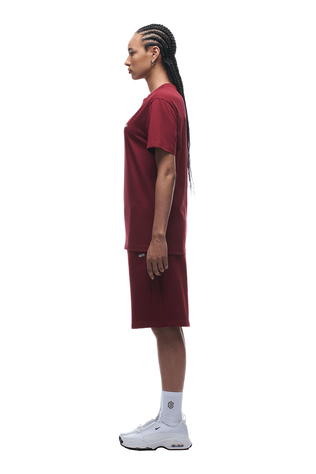 A person with long braids stands in profile against a plain green background, wearing the 6PM T-SHIRT REGULAR FIT WINE RED with matching shorts, white socks, and white sneakers.