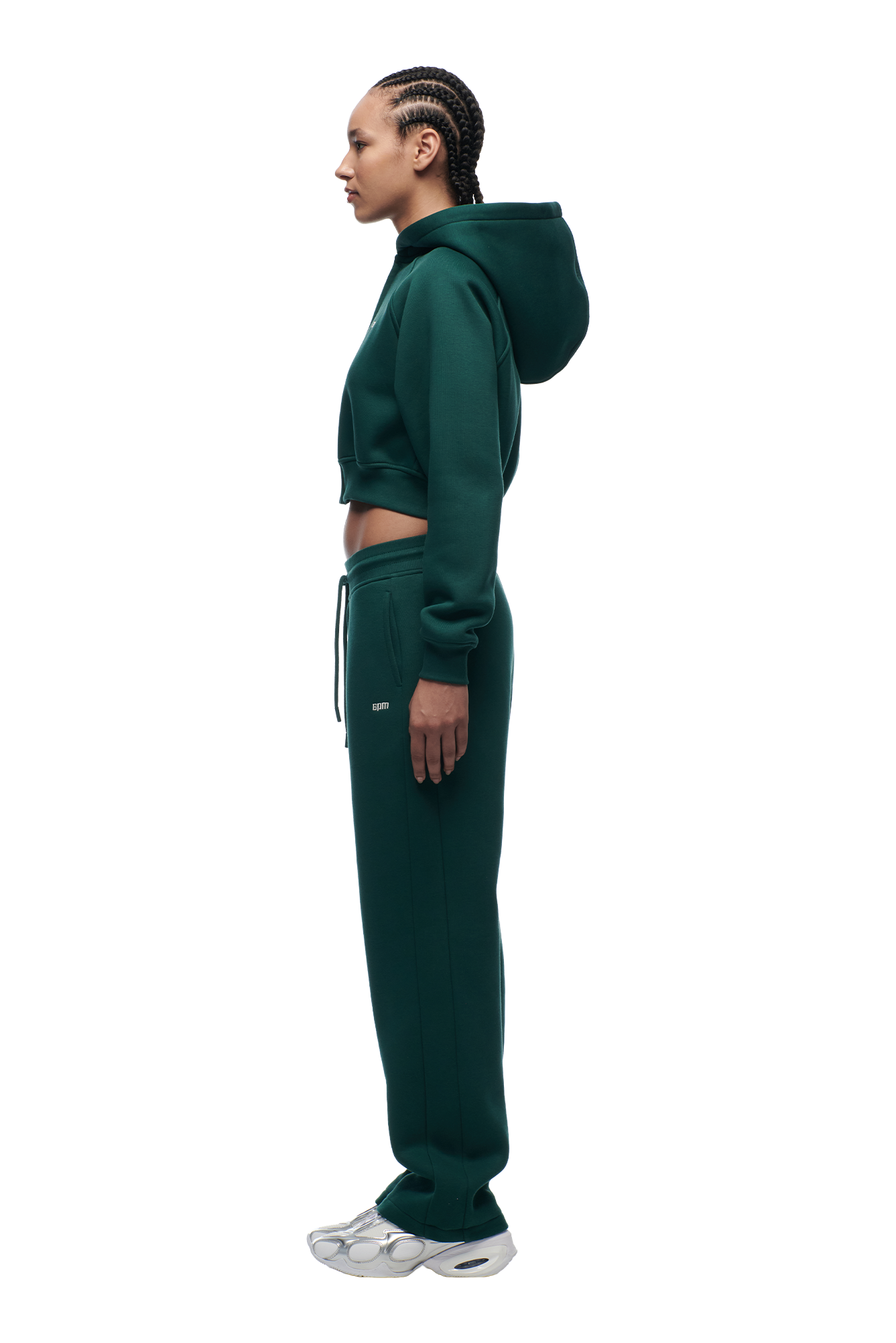 A person with braided hair stands in profile, wearing the 6PM WMNS Cropped Zip Hoodie Evergreen, matching sweatpants, and white sneakers against a plain white background.