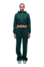A woman with braided hair faces forward against a white background, wearing the 6PM WMNS CROPPED ZIP HOODIE EVERGREEN and matching sweatpants, paired with white sneakers.