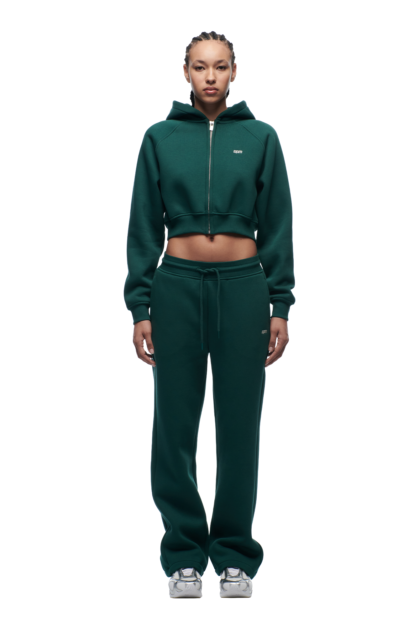 A woman with braided hair faces forward against a white background, wearing the 6PM WMNS CROPPED ZIP HOODIE EVERGREEN and matching sweatpants, paired with white sneakers.