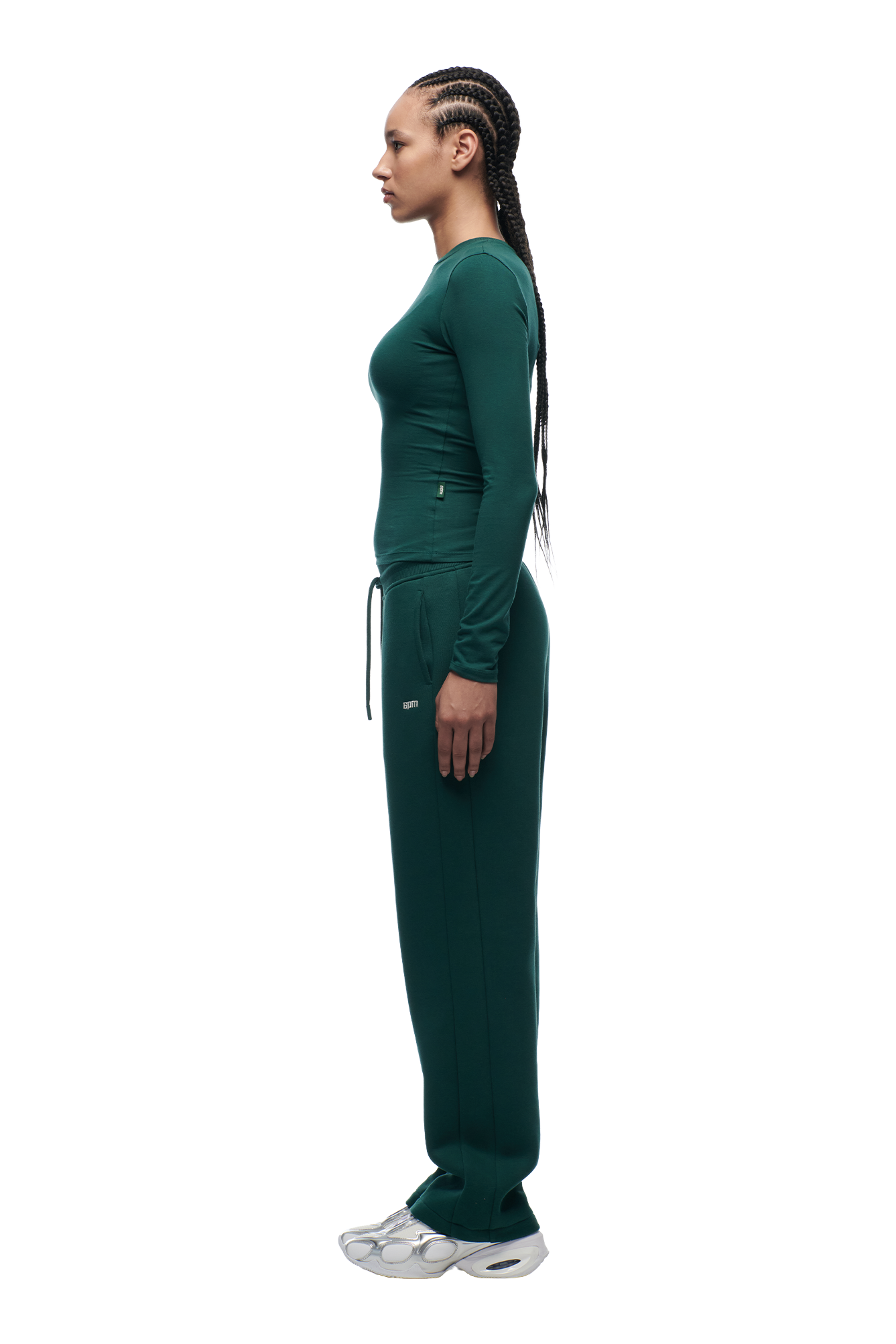 A woman with long braided hair poses in profile against a white background, wearing the 6PM WMNS LONGSLEEVE EVERGREEN top with matching pants and silver sneakers.