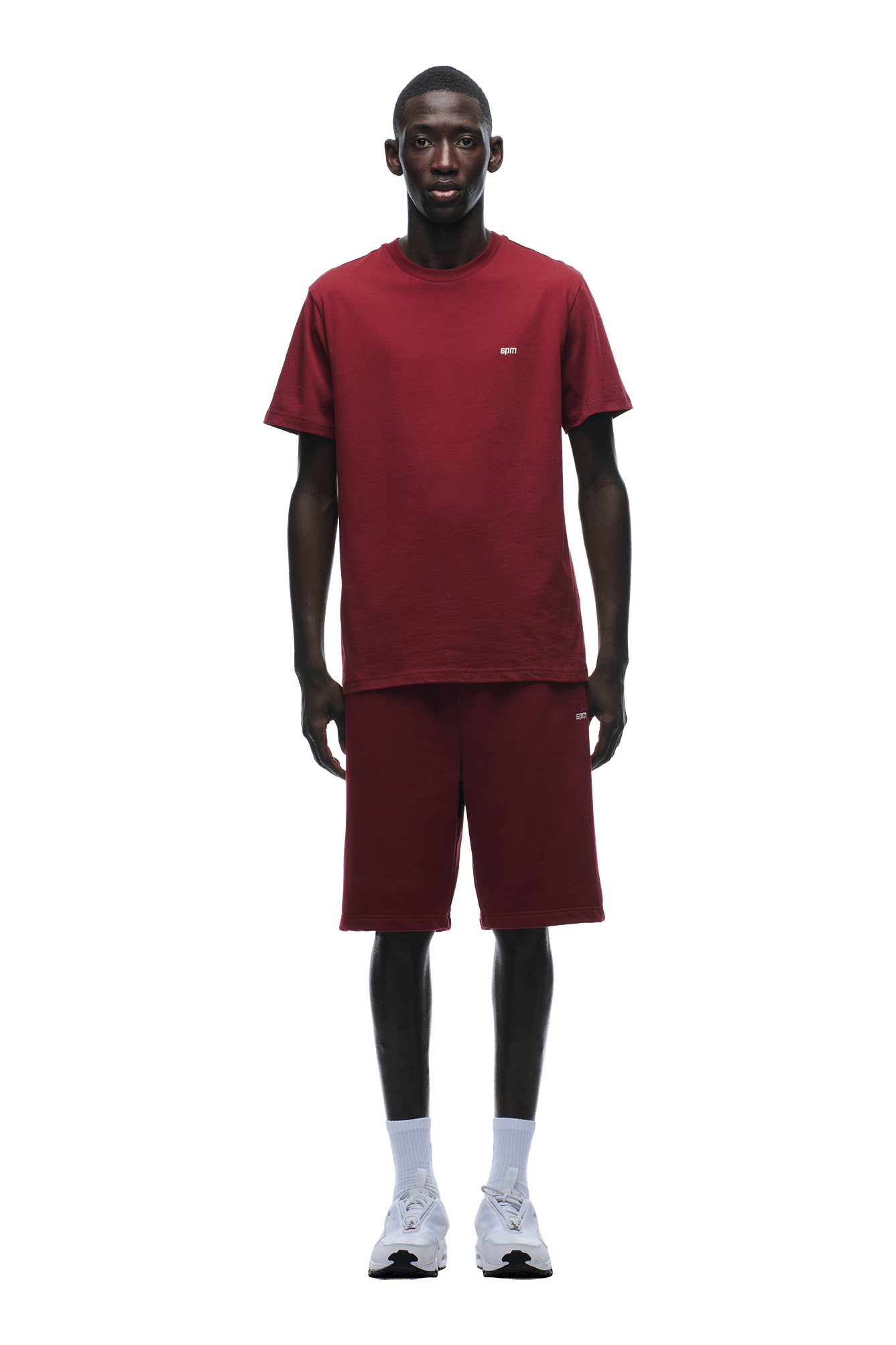 A man stands facing forward in a matching 6PM T-SHIRT REGULAR FIT WINE RED and shorts set, paired with white socks and sneakers, showing how a simple look can stand out against a green background.