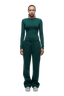 A woman with braided hair models the 6PM WMNS LONGSLEEVE EVERGREEN top and matching drawstring sweatpants, paired with silver sneakers, standing against a plain white background.