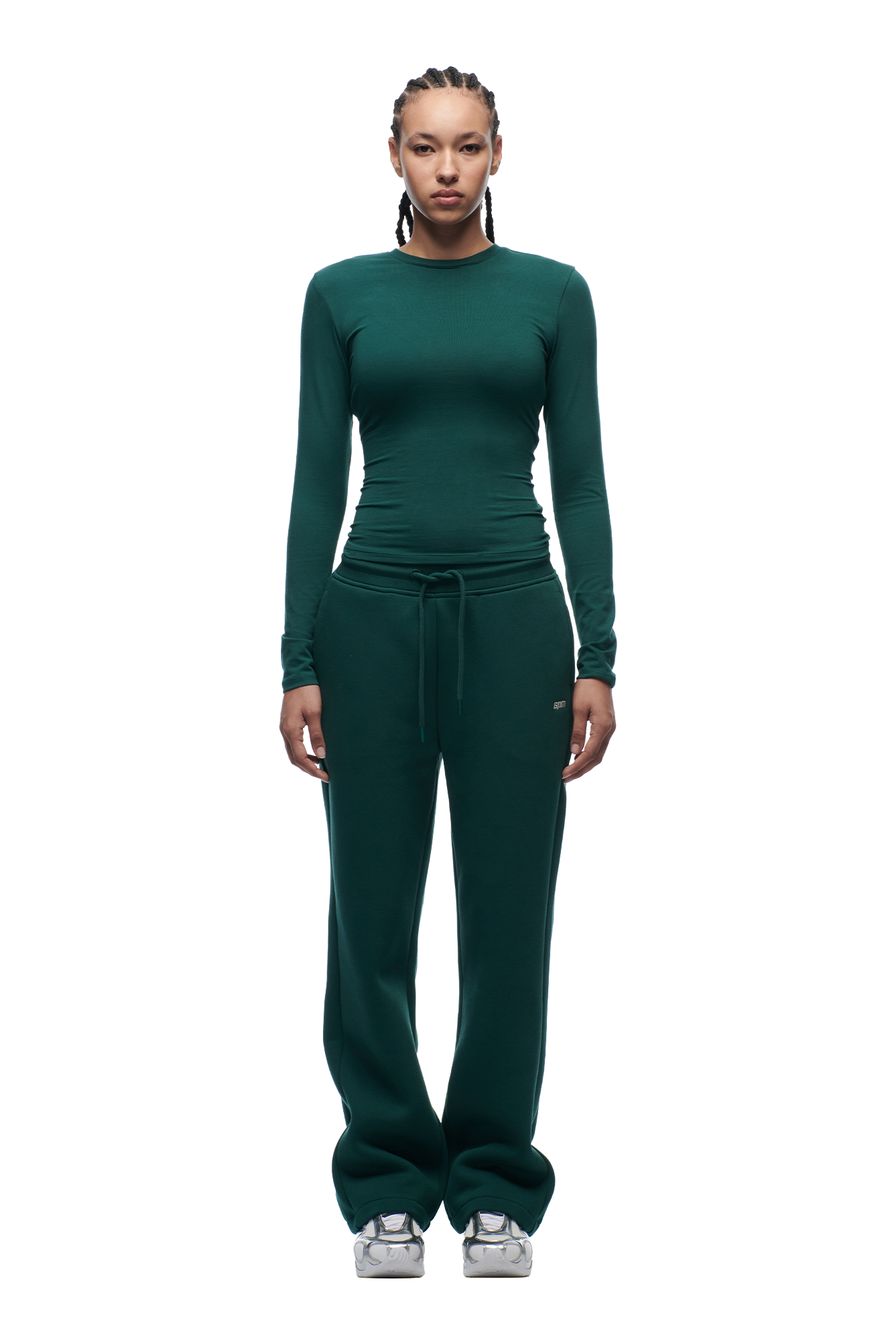 A woman with braided hair models the 6PM WMNS LONGSLEEVE EVERGREEN top and matching drawstring sweatpants, paired with silver sneakers, standing against a plain white background.