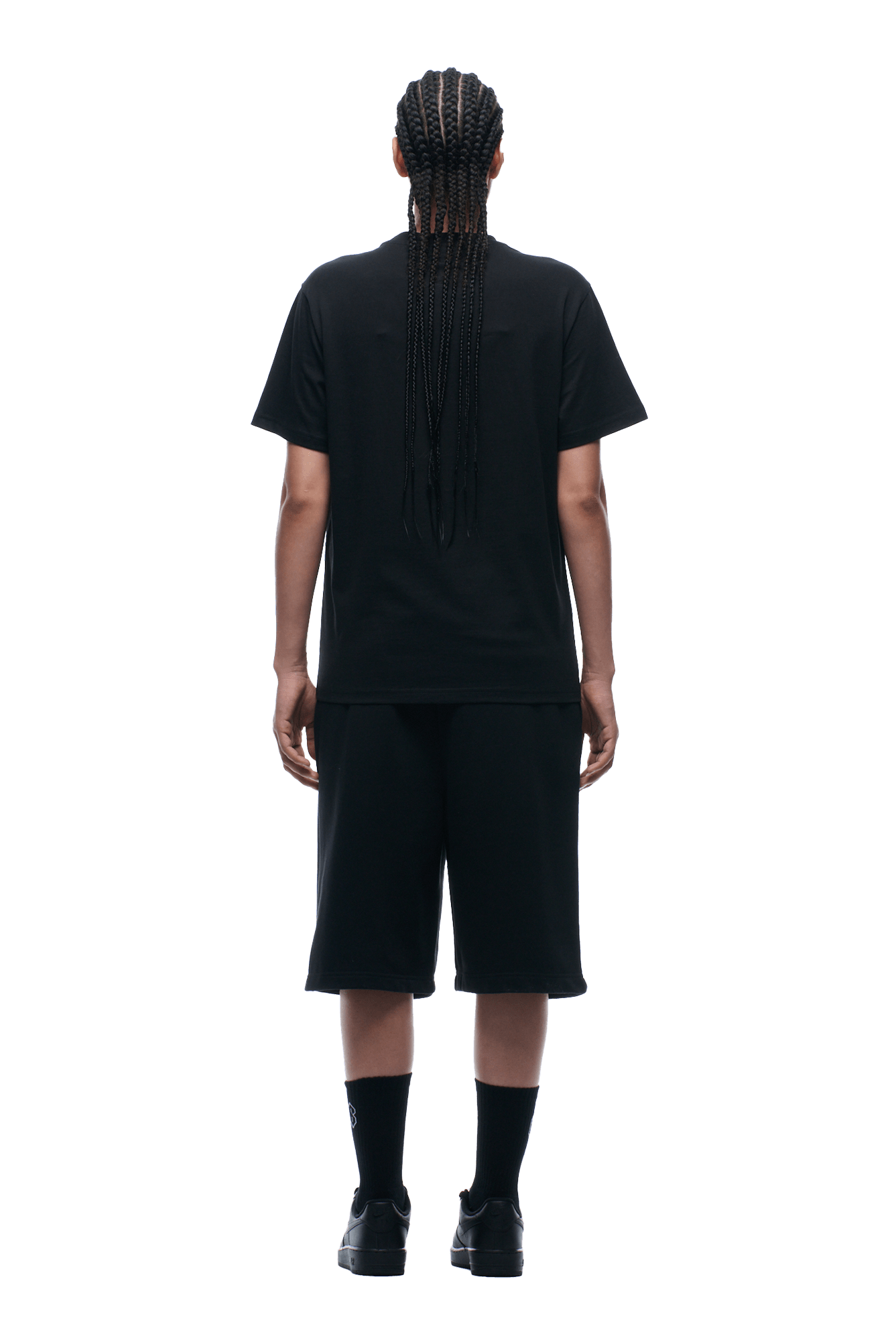 A person with long braids wears the 6PM GRAN TURISMO TEE BLACK, paired with black shorts, socks, and shoes. They stand facing away from the camera against a transparent background.