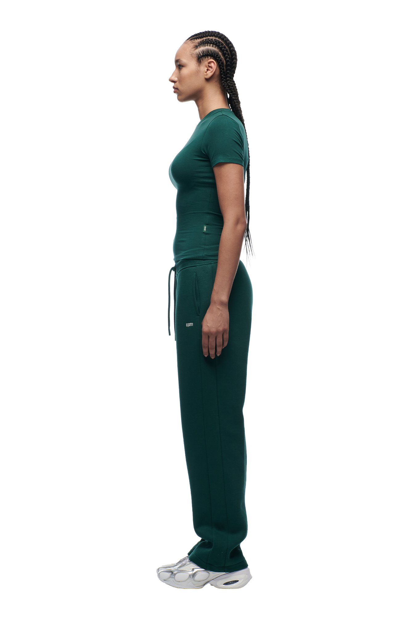 A person with long braided hair stands in profile, wearing the 6PM WMNS T-SHIRT EVERGREEN and matching green drawstring sweatpants with gray sneakers, against a plain white background.