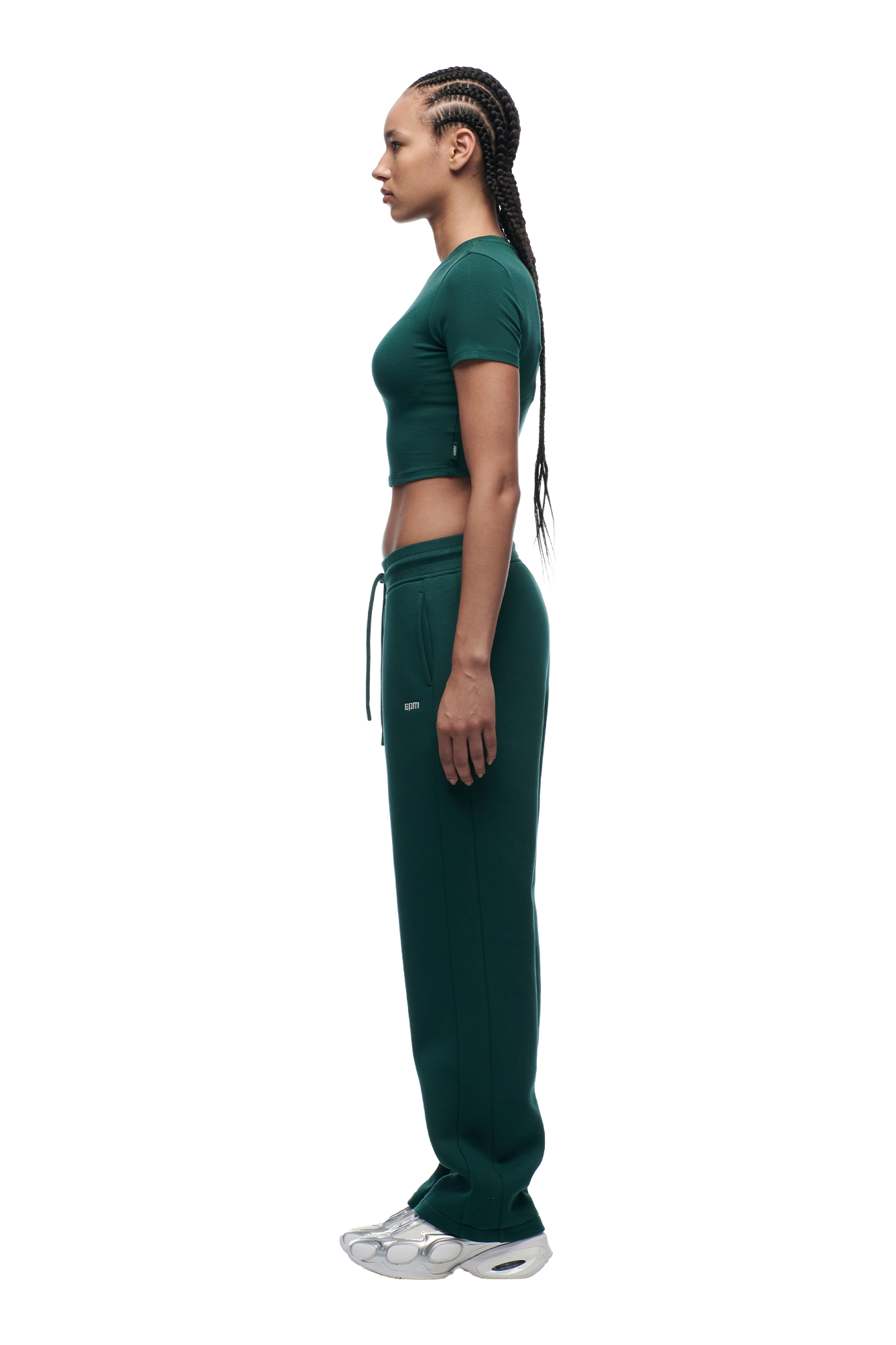 A woman with long braided hair stands in profile, wearing the 6PM WMNS Cropped T-Shirt Evergreen and matching green sweatpants with silver sneakers against a plain white background.