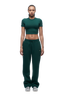 A woman with braided hair wears the 6PM WMNS CROPPED T-SHIRT EVERGREEN and matching green sweatpants with drawstrings, paired with silver sneakers, standing against a white background.