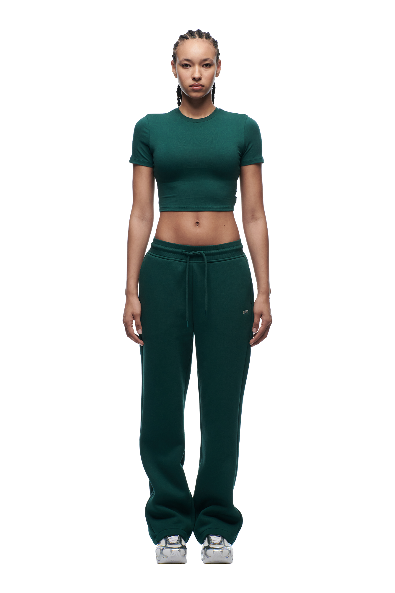 A woman with braided hair wears the 6PM WMNS CROPPED T-SHIRT EVERGREEN and matching green sweatpants with drawstrings, paired with silver sneakers, standing against a white background.