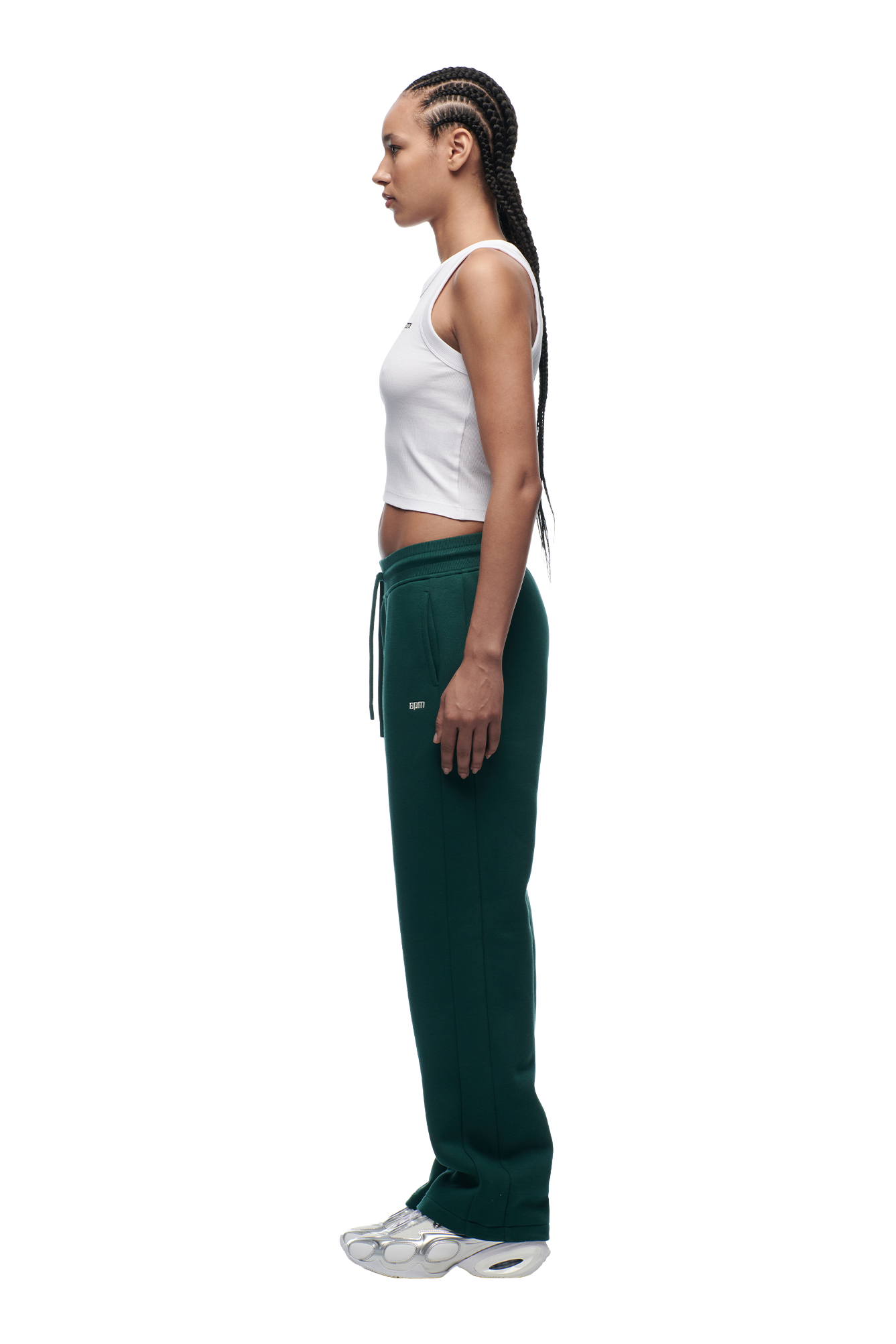 A woman with braided hair stands in profile wearing the WMNS OPEN LEG EVERGREEN green drawstring sweatpants by 6PM, a white sleeveless crop top, and white sneakers against a plain white background.