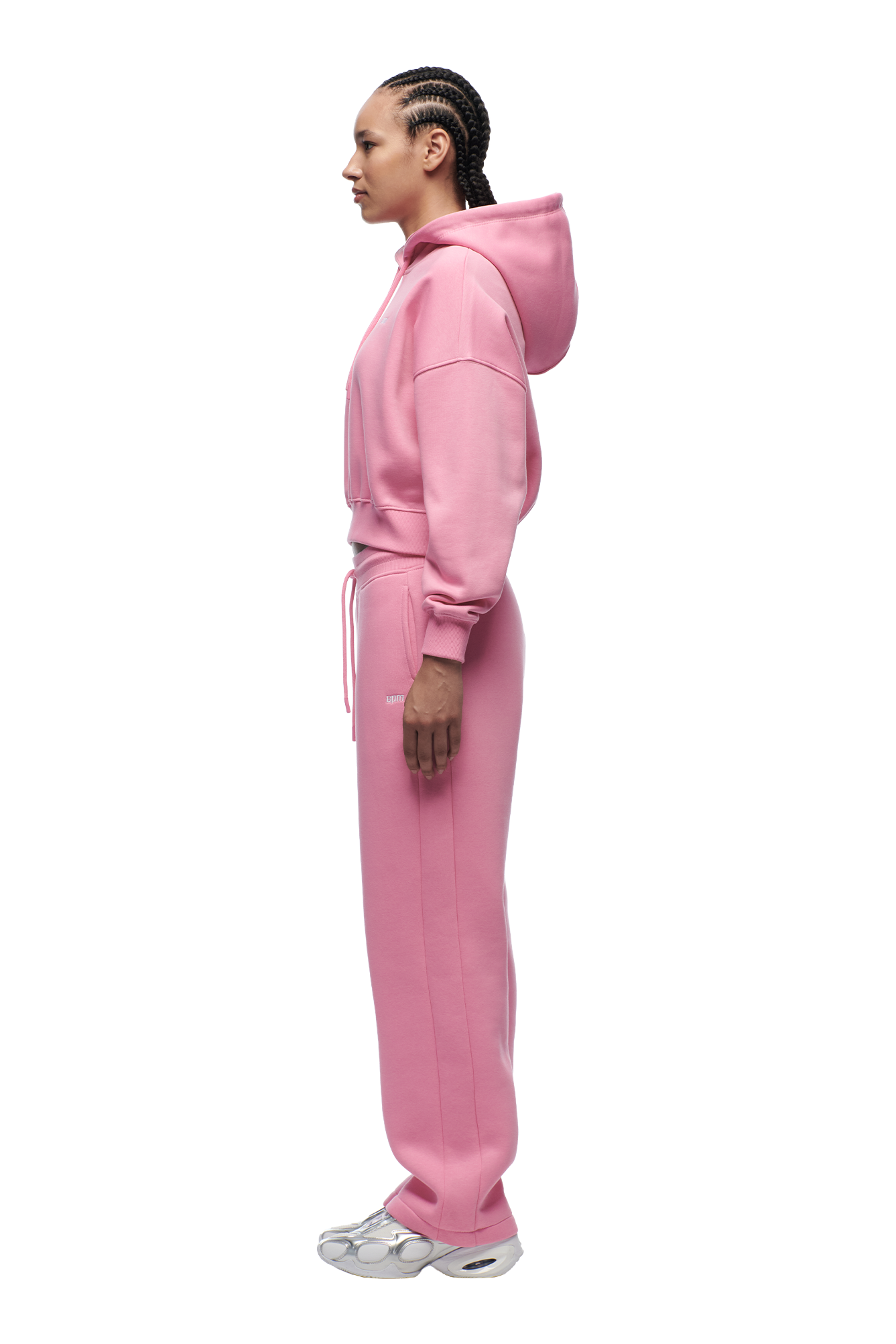 A person with braided hair stands in profile, wearing the 6PM WMNS CROPPED HOODIE BUBBLEGLOW in pink with matching sweatpants and white sneakers, set against a plain white background.
