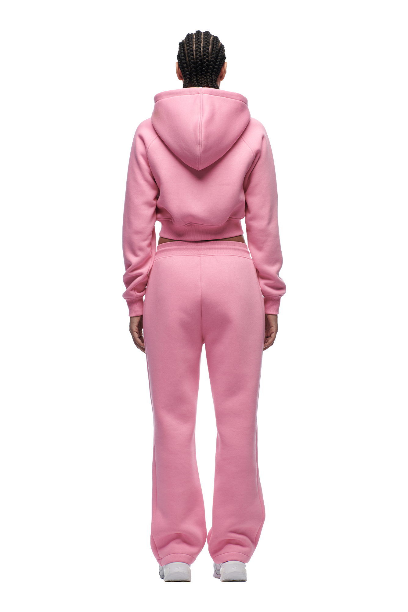 A person with braided hair is seen from the back wearing the 6PM WMNS CROPPED ZIP HOODIE BUBBLEGLOW in pink and matching sweatpants, paired with white sneakers for a casual, comfortable look.