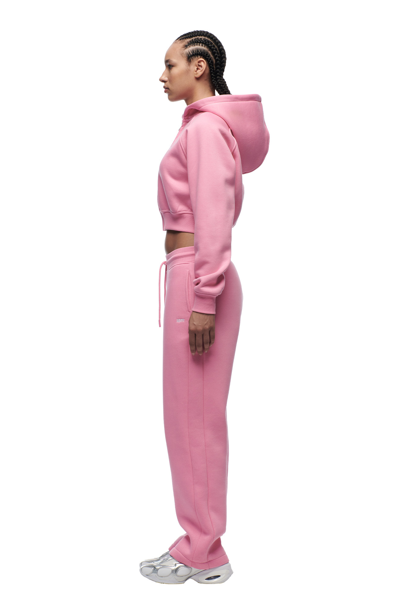 A person stands in profile wearing the 6PM WMNS CROPPED ZIP HOODIE BUBBLEGLOW and matching sweatpants with white sneakers against a plain white background.