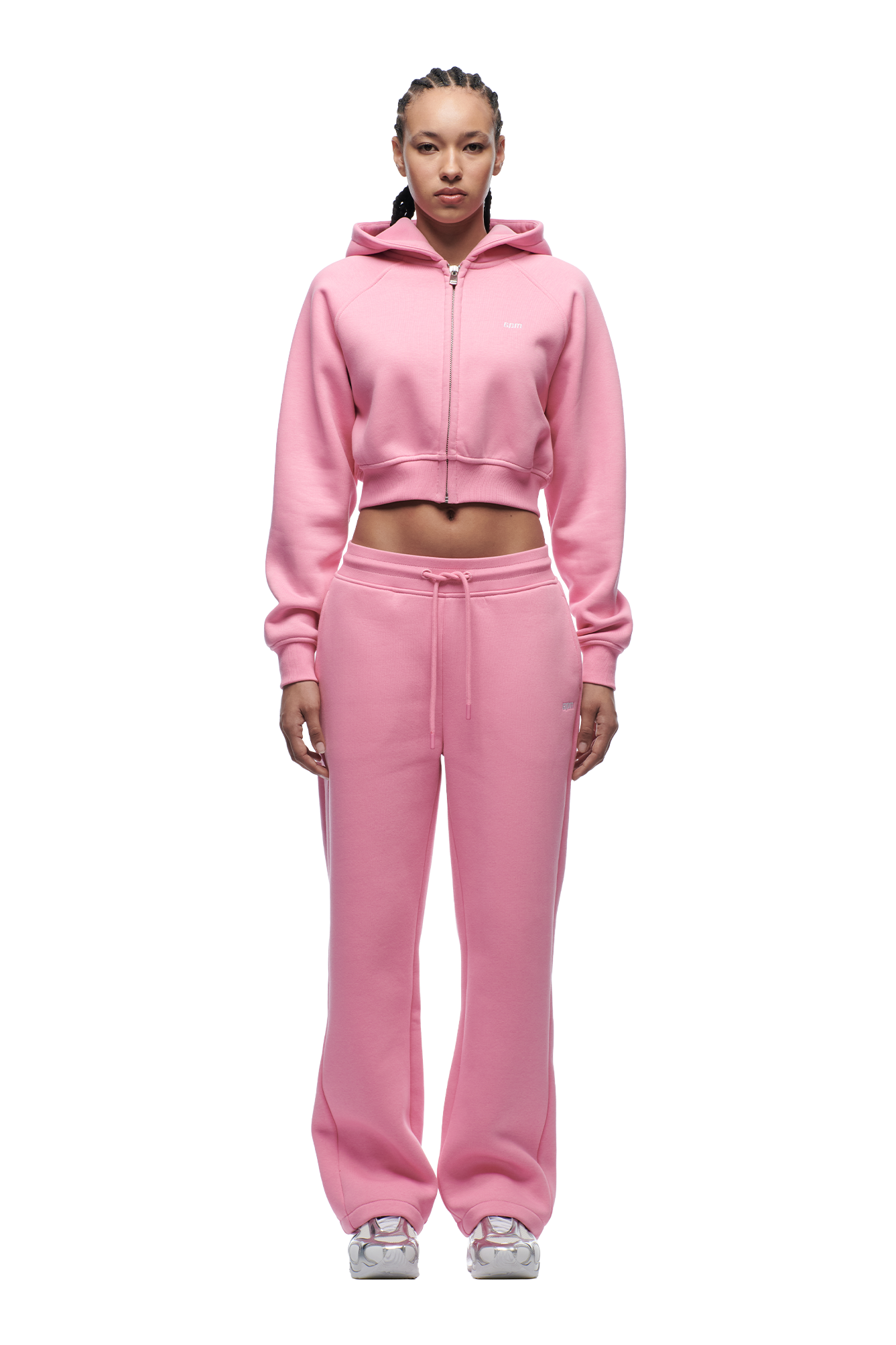 A woman with braided hair faces forward, wearing the 6PM WMNS CROPPED ZIP HOODIE BUBBLEGLOW in light pink, matching sweatpants, and white sneakers against a plain white background.