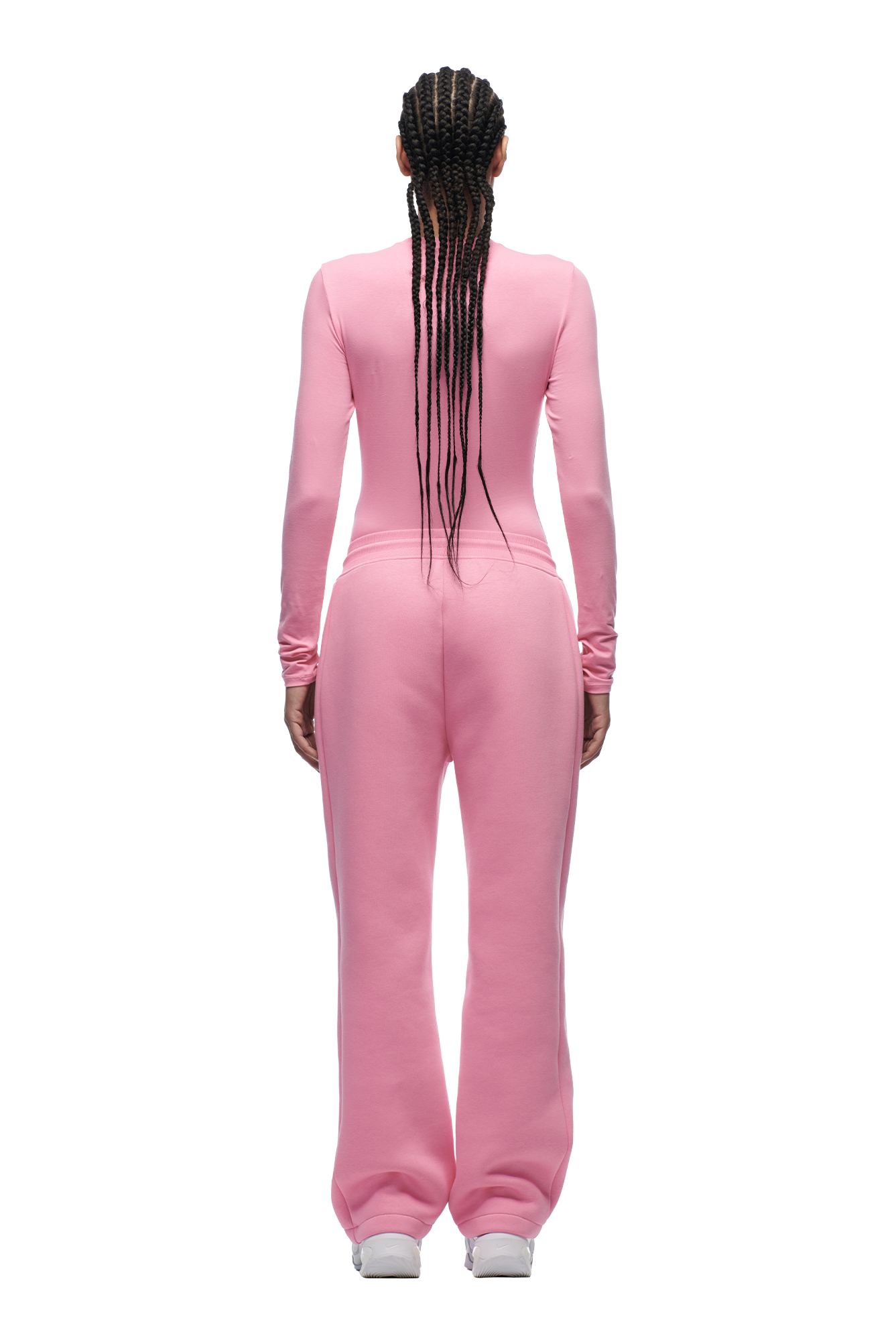 A person with long braids faces away, wearing the 6PM WMNS LONGSLEEVE BUBBLEGLOW pink top and loose pink sweatpants with white sneakers, set against a plain white background.