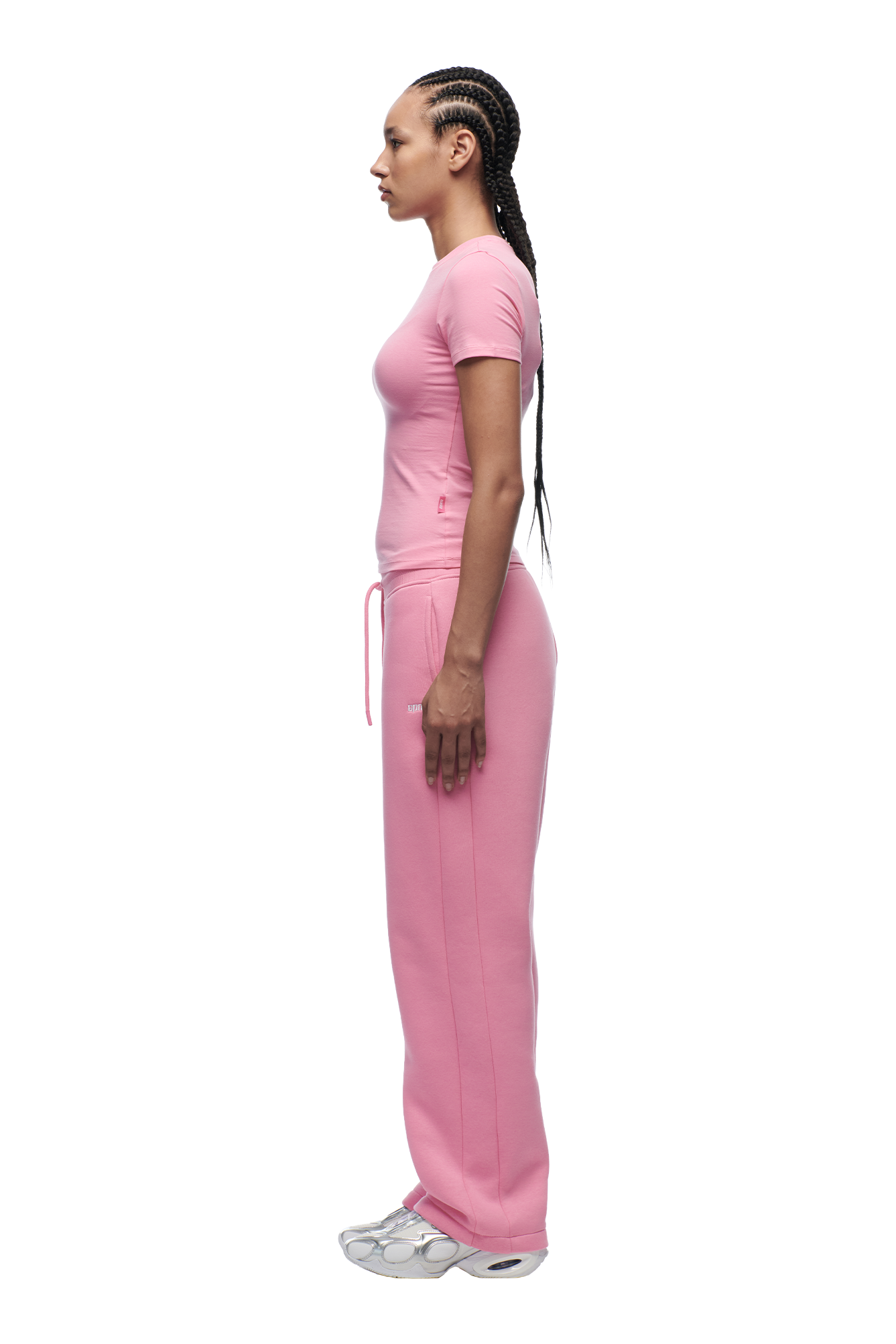 A woman with long braided hair wears the 6PM WMNS T-SHIRT BUBBLEGLOW, matching pink sweatpants, and white sneakers as she stands in profile against a plain white background.