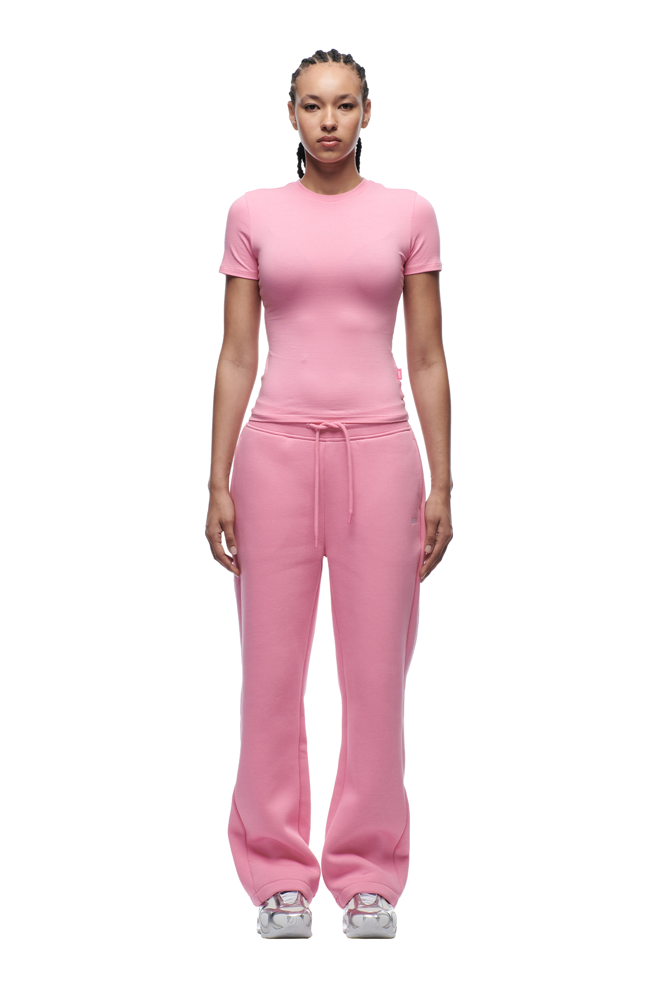 A person with braided hair faces forward, wearing the 6PM WMNS T-SHIRT BUBBLEGLOW, loose pink sweatpants with a drawstring, and white sneakers against a plain white background.