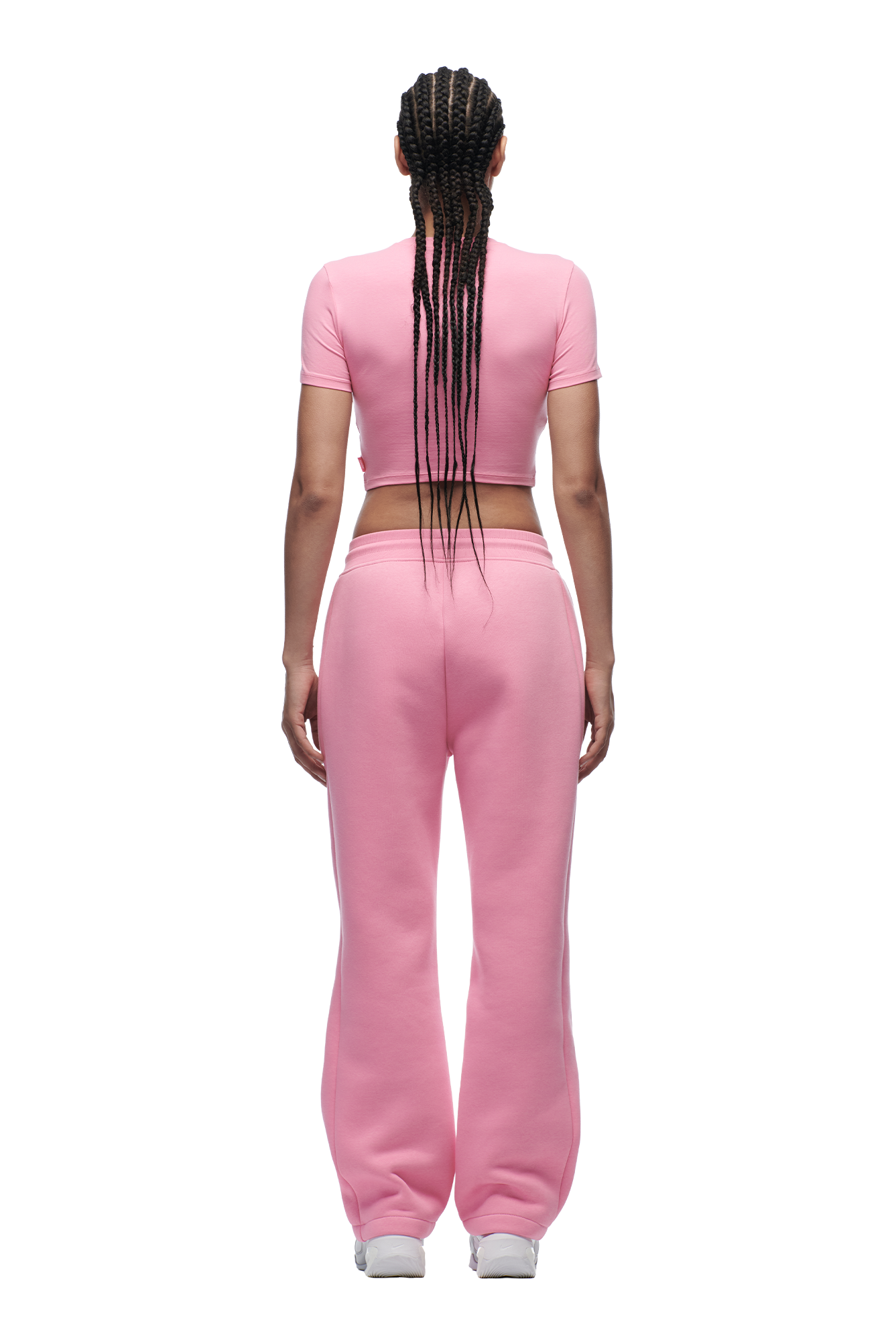 A woman with braided hair stands against a plain white background wearing the 6PM WMNS CROPPED T-SHIRT BUBBLEGLOW, matching pink sweatpants, and white sneakers.