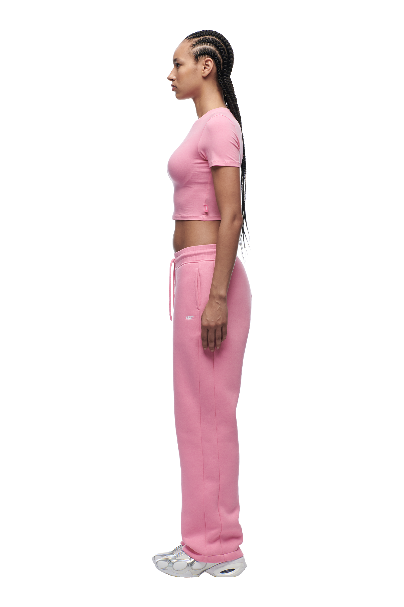 A woman with long braids stands in profile against a plain white background, wearing the 6PM WMNS CROPPED T-SHIRT BUBBLEGLOW and matching pink sweatpants, paired with silver sneakers.