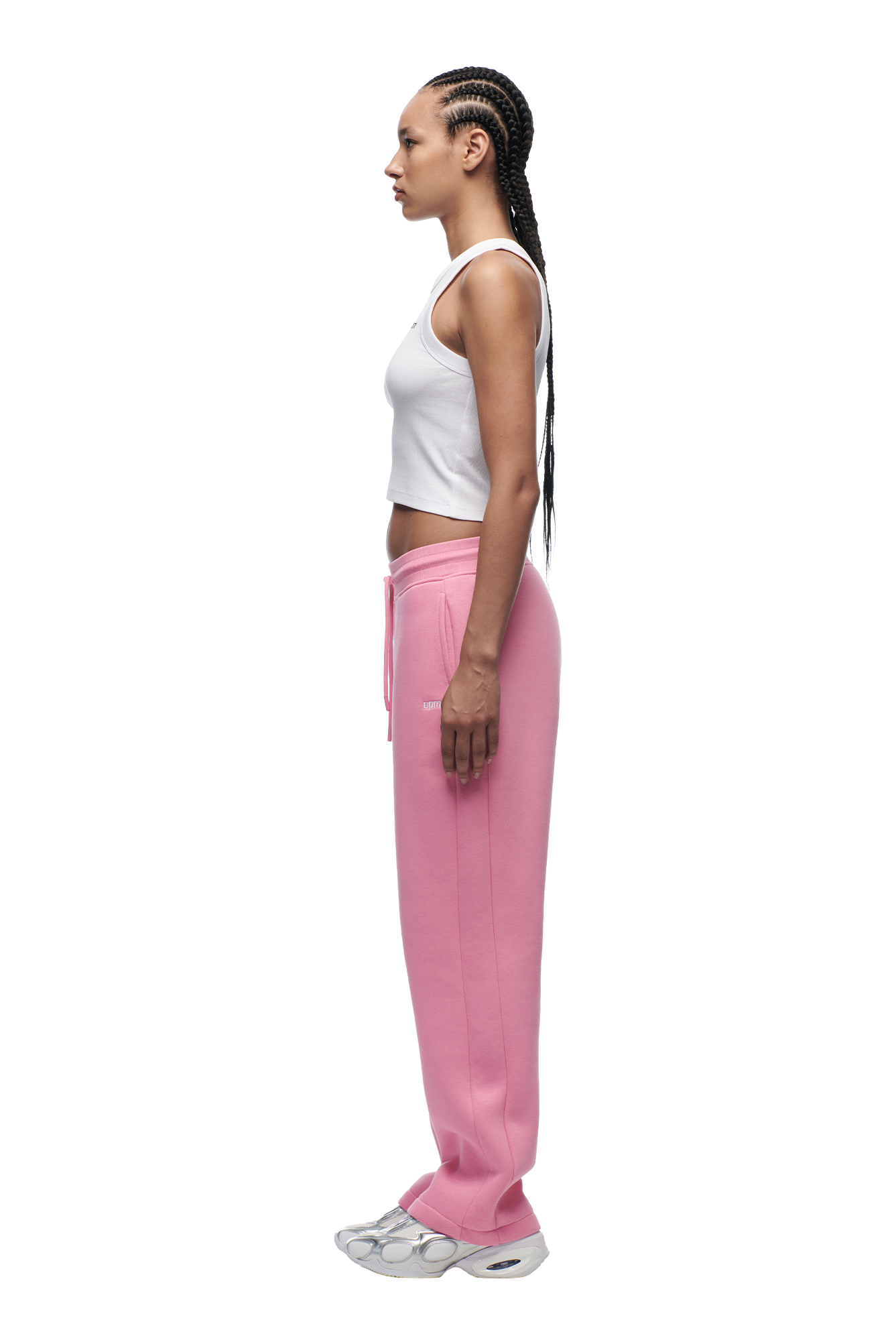 A woman with long braided hair stands in profile wearing 6PM's WMNS OPEN LEG BUBBLEGLOW pink sweatpants, a white sleeveless crop top, and gray sneakers against a plain white background.