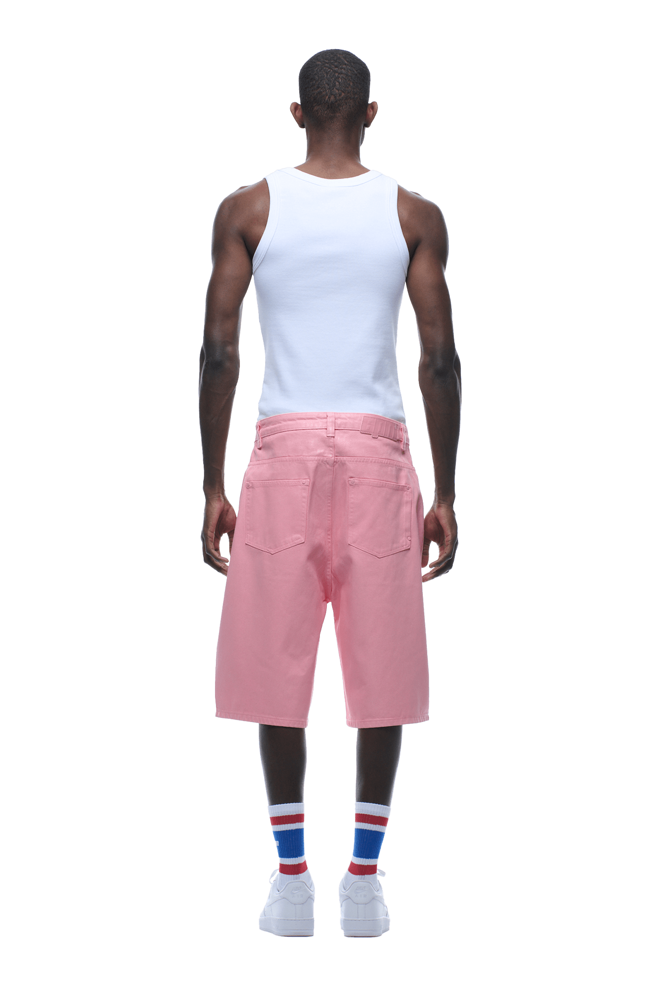 A man stands facing away in a white tank top, COATED DENIM SHORTS PINK by 6PM, white socks with red and blue stripes, and white sneakers against a plain background.