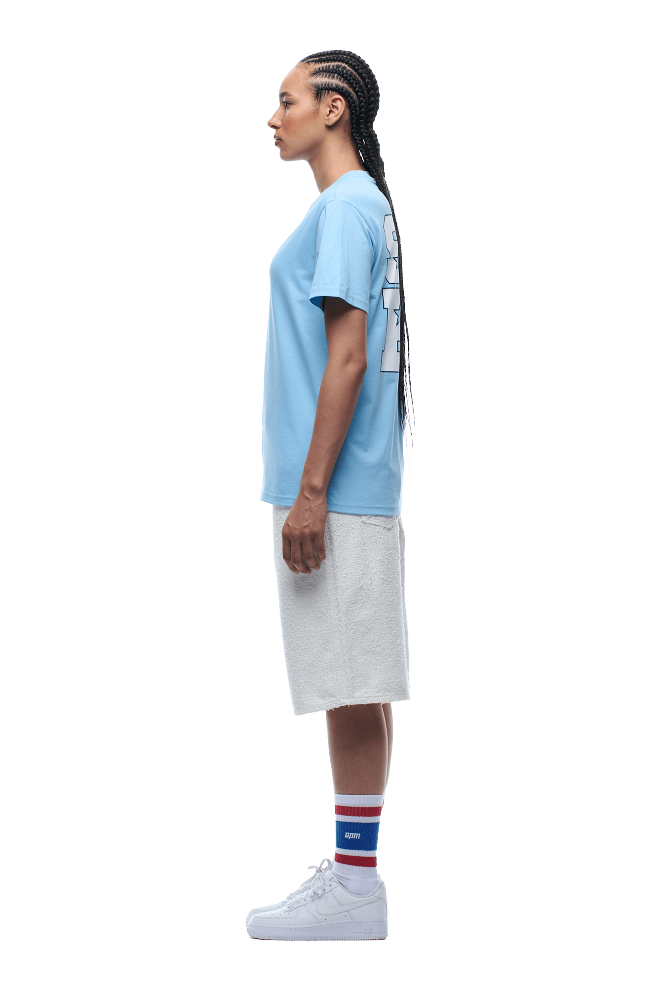 A person with long braided hair and a neutral expression stands in profile wearing the 6PM COLLEGE STAR TEE BLUE, white knee-length shorts, white sneakers, and blue, red, and white striped socks.
