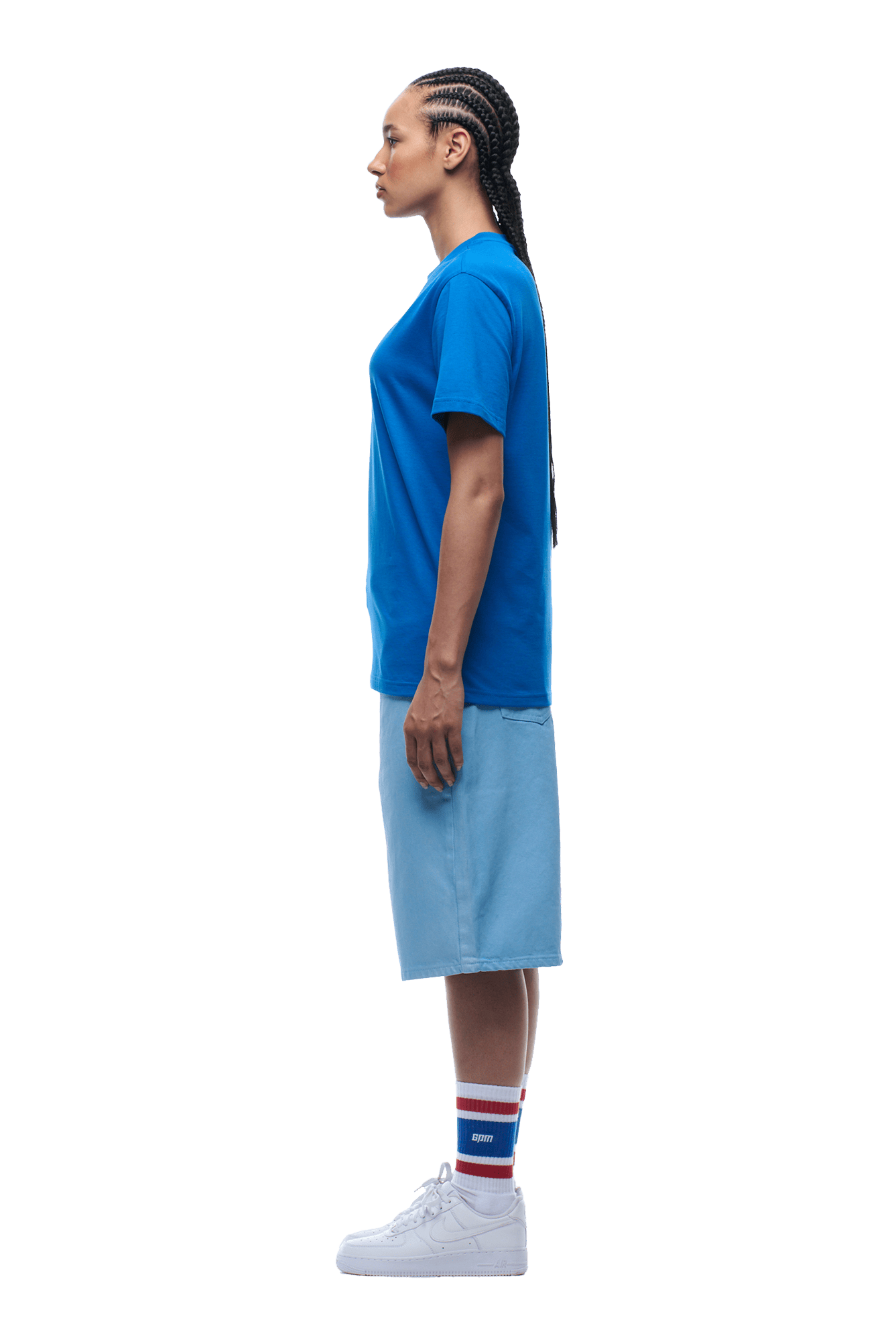 A person with braided hair stands in profile on a transparent background, wearing the 6PM APPLE BOY TEE BLUE, light blue knee-length shorts, white sneakers, and blue, white, and red striped socks.
