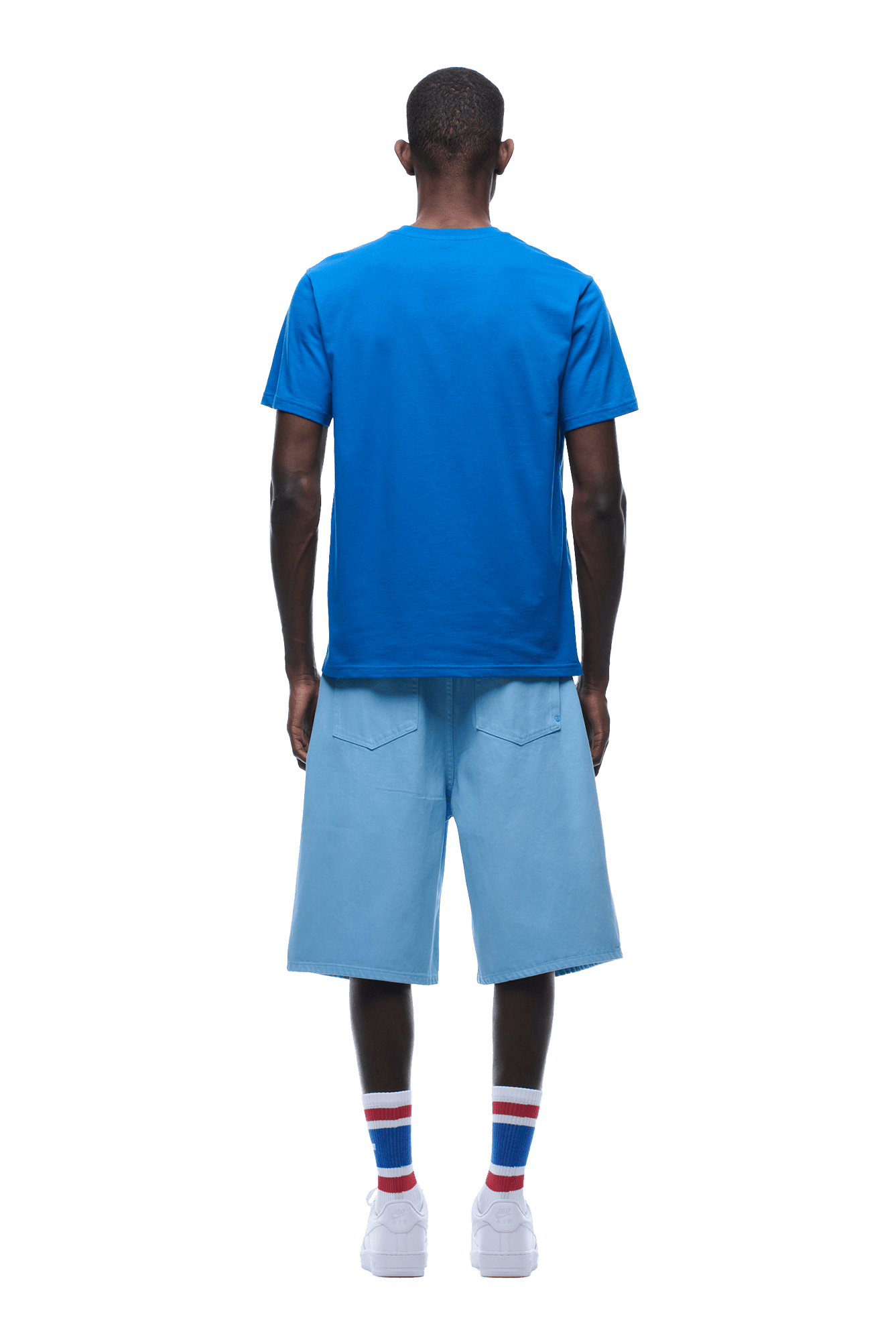 A man stands facing away, wearing the 6PM APPLE BOY TEE BLUE, light blue knee-length shorts, red-white-blue striped socks, and white sneakers on a transparent background.
