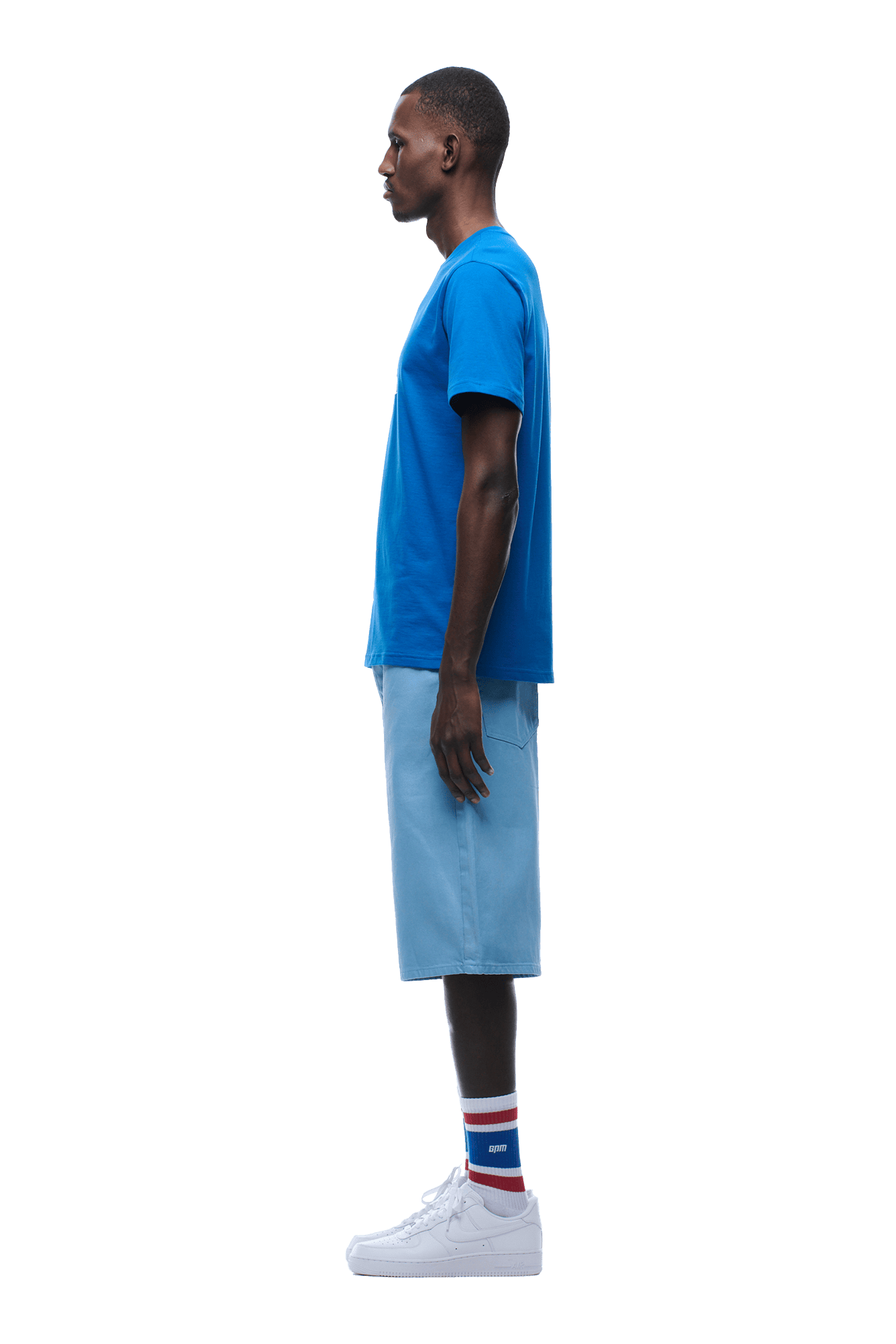 A man stands in profile facing left, wearing 6PMs APPLE BOY TEE BLUE, light blue shorts, white sneakers, and white socks with blue and red stripes. The background is transparent.