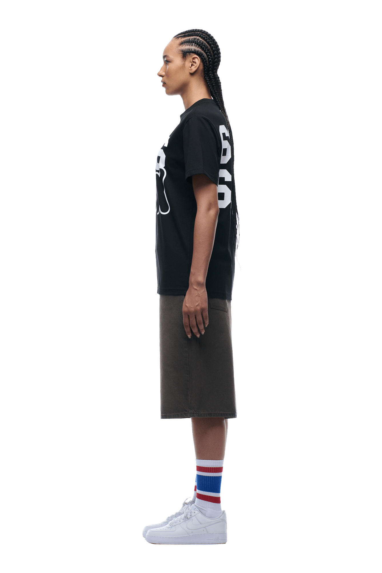 A person with braided hair stands in profile on a plain background, wearing the 6PM 660660 TEE BLACK, a dark knee-length skirt, striped socks, and white sneakers.