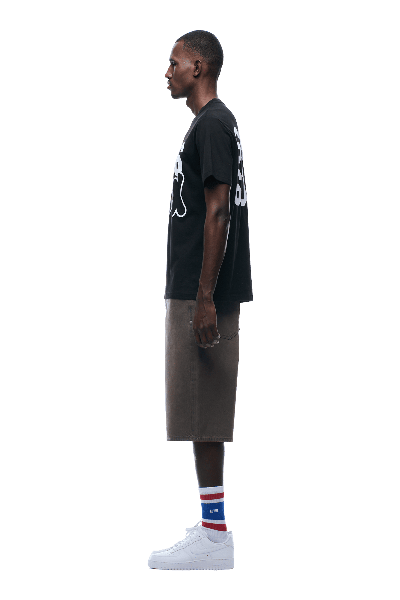 A man stands in profile, wearing the 6PM 660660 TEE BLACK, knee-length brown shorts, white sneakers, and white socks with blue and red stripes, against a transparent background.