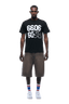 A man faces forward wearing the 6PM 660660 TEE BLACK, long brown shorts, white sneakers, and striped socks; background is transparent.
