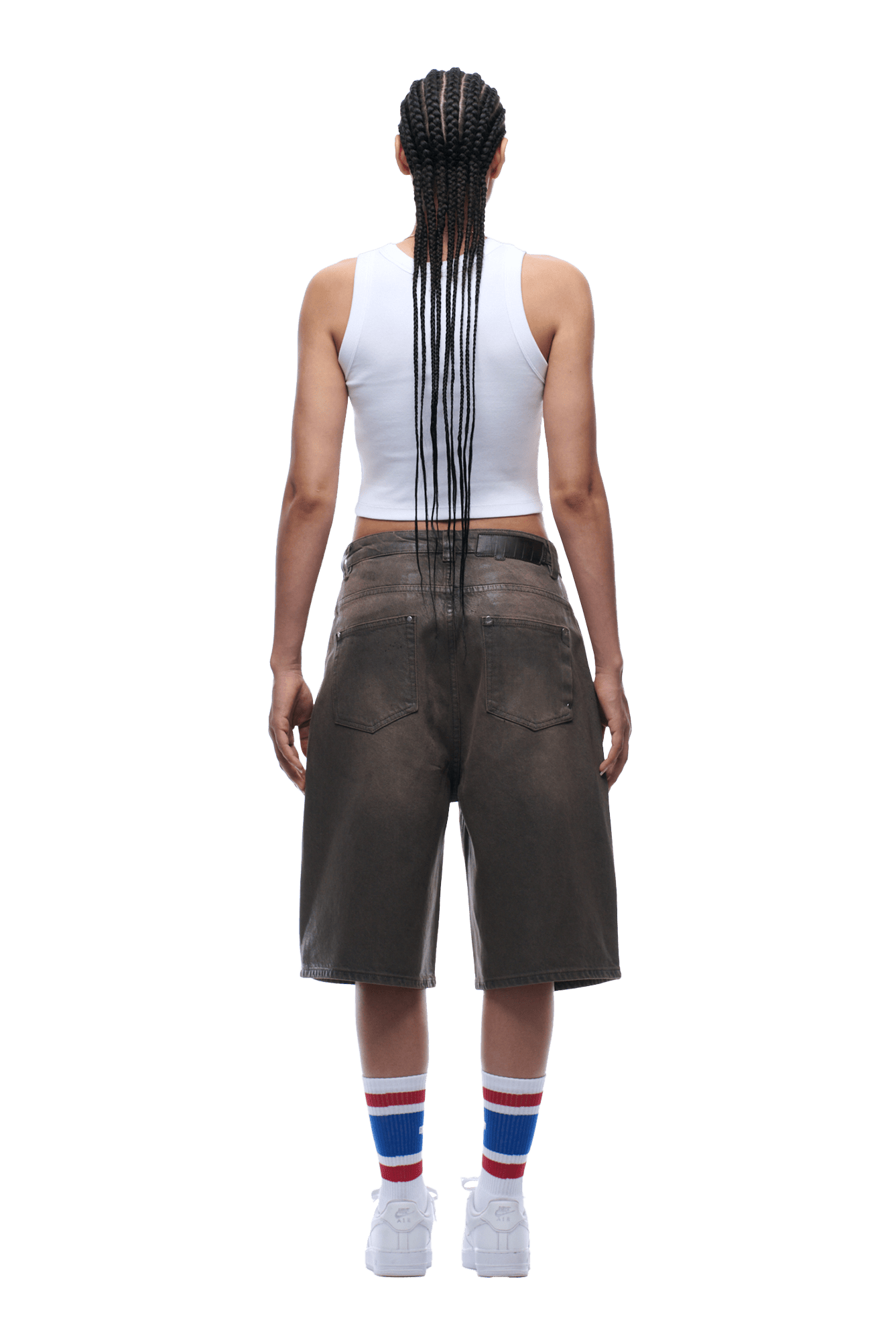 A person with long braided hair, facing away, wears a white tank top, 6PM COATED DENIM SHORTS WASHED BROWN, white sneakers, and white socks with red and blue stripes.