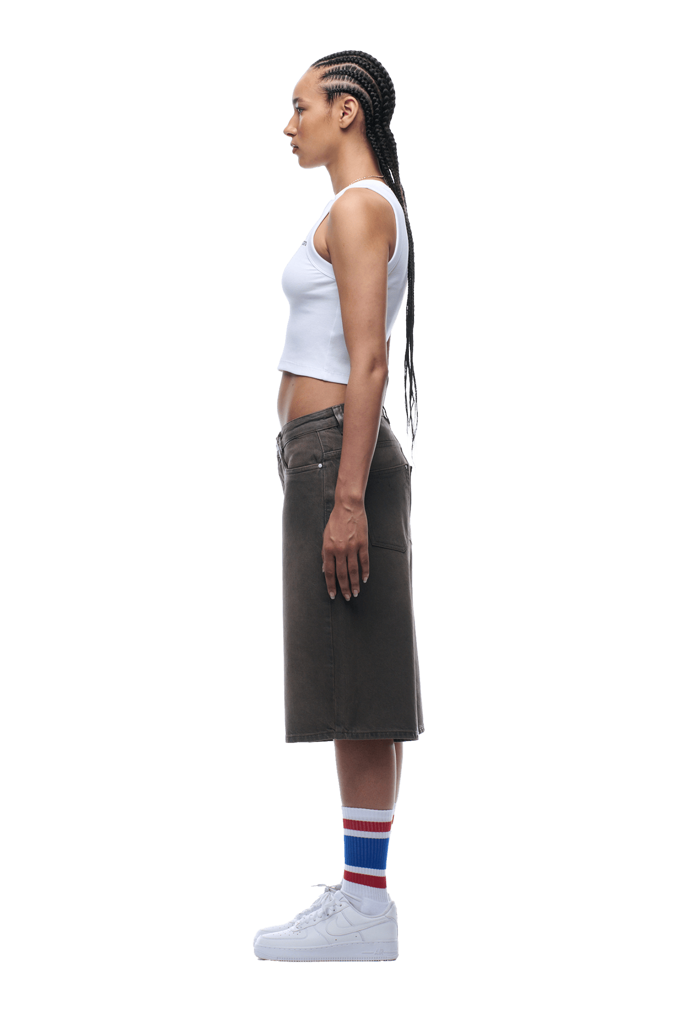 A woman with long braids stands in profile, wearing a white crop top, COATED DENIM SHORTS WASHED BROWN from 6PM, striped socks, and white sneakers on a transparent background.