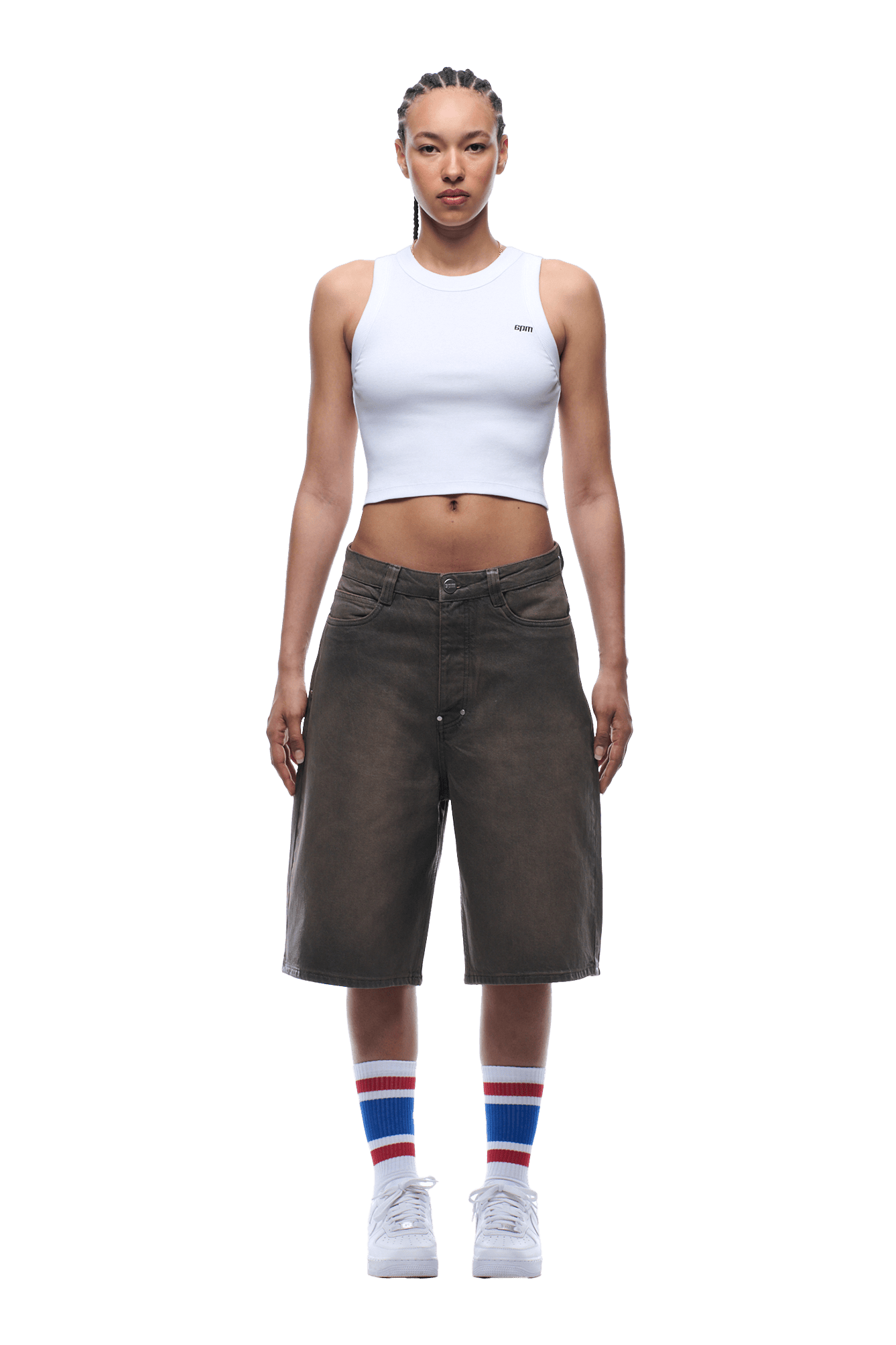 A woman with braided hair and a neutral expression faces forward, wearing 6PM COATED DENIM SHORTS WASHED BROWN, a white cropped tank top, white sneakers, and colorful striped socks.